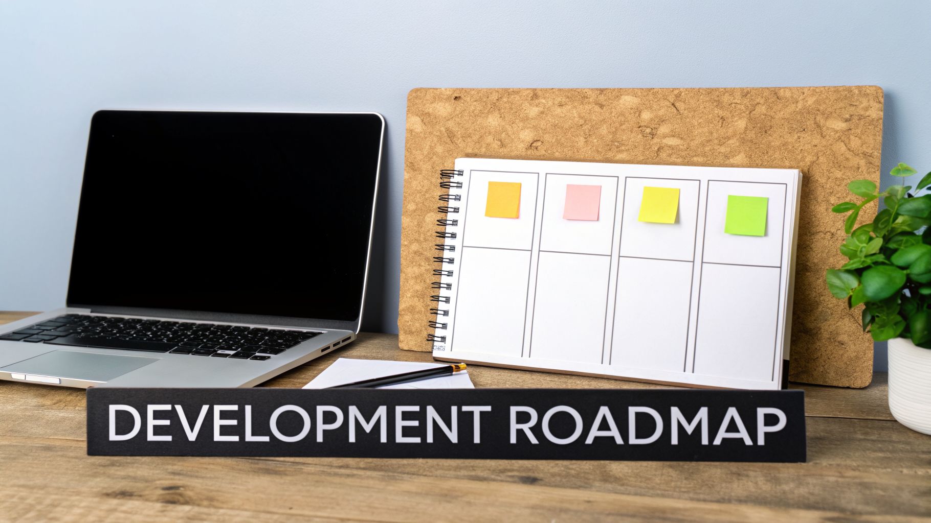 Laptop, project notebook with colorful sticky notes, and a 'DEVELOPMENT ROADMAP' sign on a wooden desk.