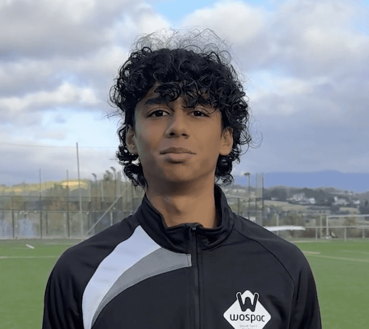 Person with tattoos and cross necklace standing by water, another figure on rocks in background.