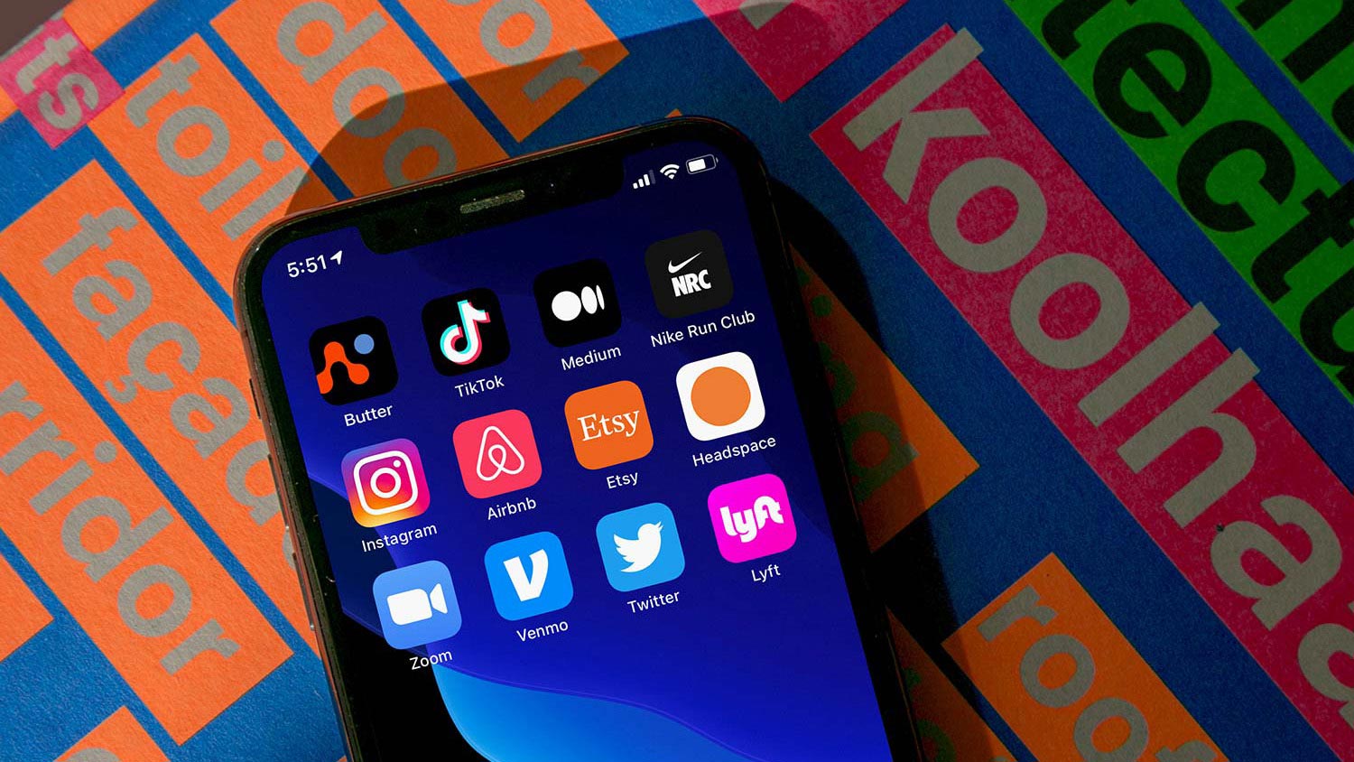 A black smartphone is resting on a surface with a repeating pattern of orange and blue squares. The screen displays several app icons, including those for TikTok, Medium, Nike Run Club, Instagram, Airbnb, Etsy, Headspace, Lyft, Zoom, Twitter, and Lyft. The time displayed on the phone is 5:51. The background features the text "Koolhaas" in large white letters on a pink-to-green gradient.