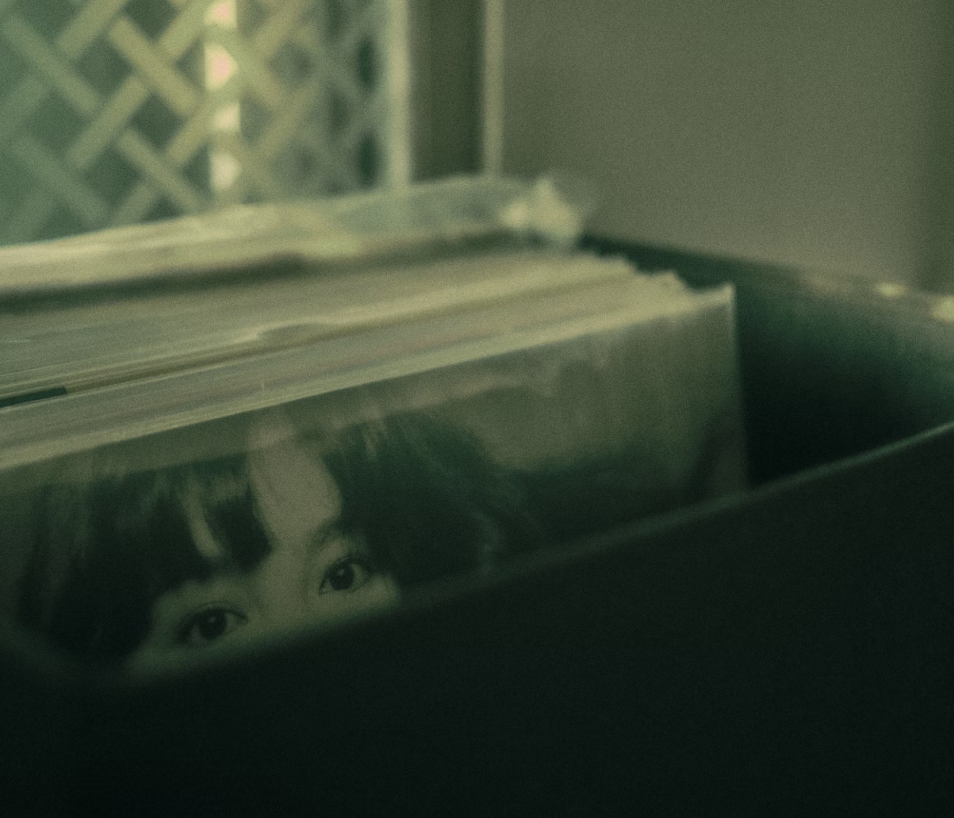 Vinyl records stored in a crate, with an album cover showing a pair of eyes visible above the stack in soft green lighting.