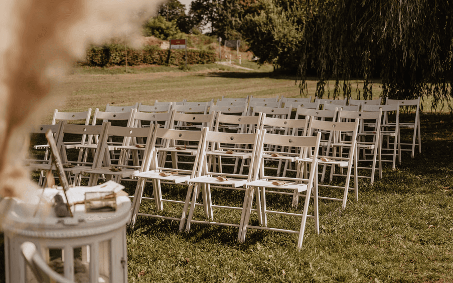 Setup für eine freie Trauung im Freien mit Reihen aus weißen Klappstühlen auf einer Wiese unter einem großen Baum