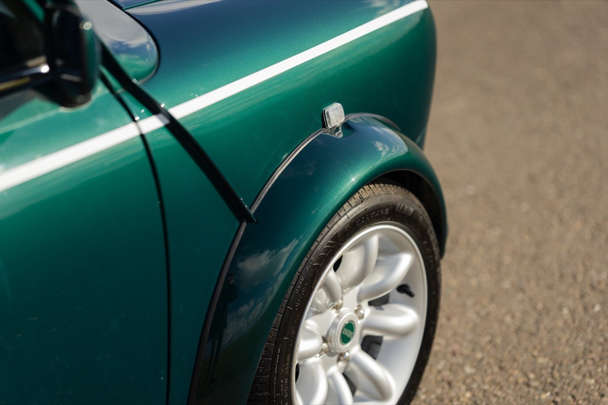 Close-up of a green car's rear fender and shiny alloy wheel on a paved surface.