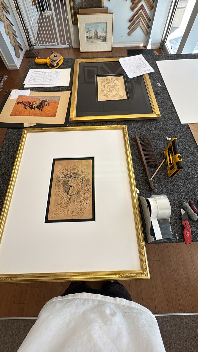 Framing studio table with a Picasso-style line drawing, a bison painting, and other artwork being prepared