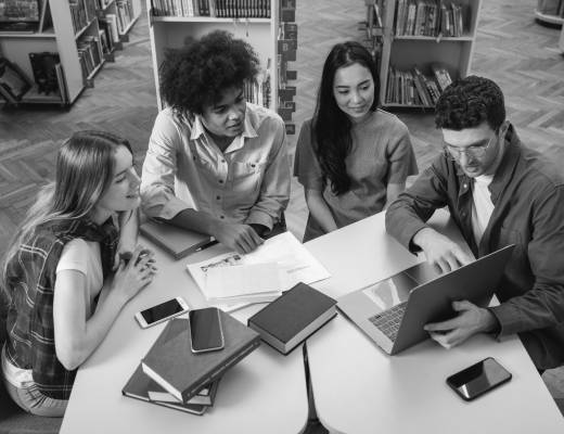 Group of four students or young professionals sitting around a table in a library, discussing and working together on a laptop with books and papers spread out.