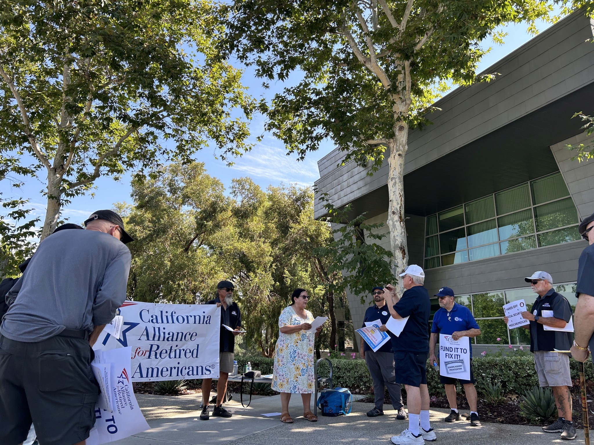 A small group gathers outside a modern building under tall trees, holding signs from the California Alliance for Retired Americans and advocating for affordable medication.