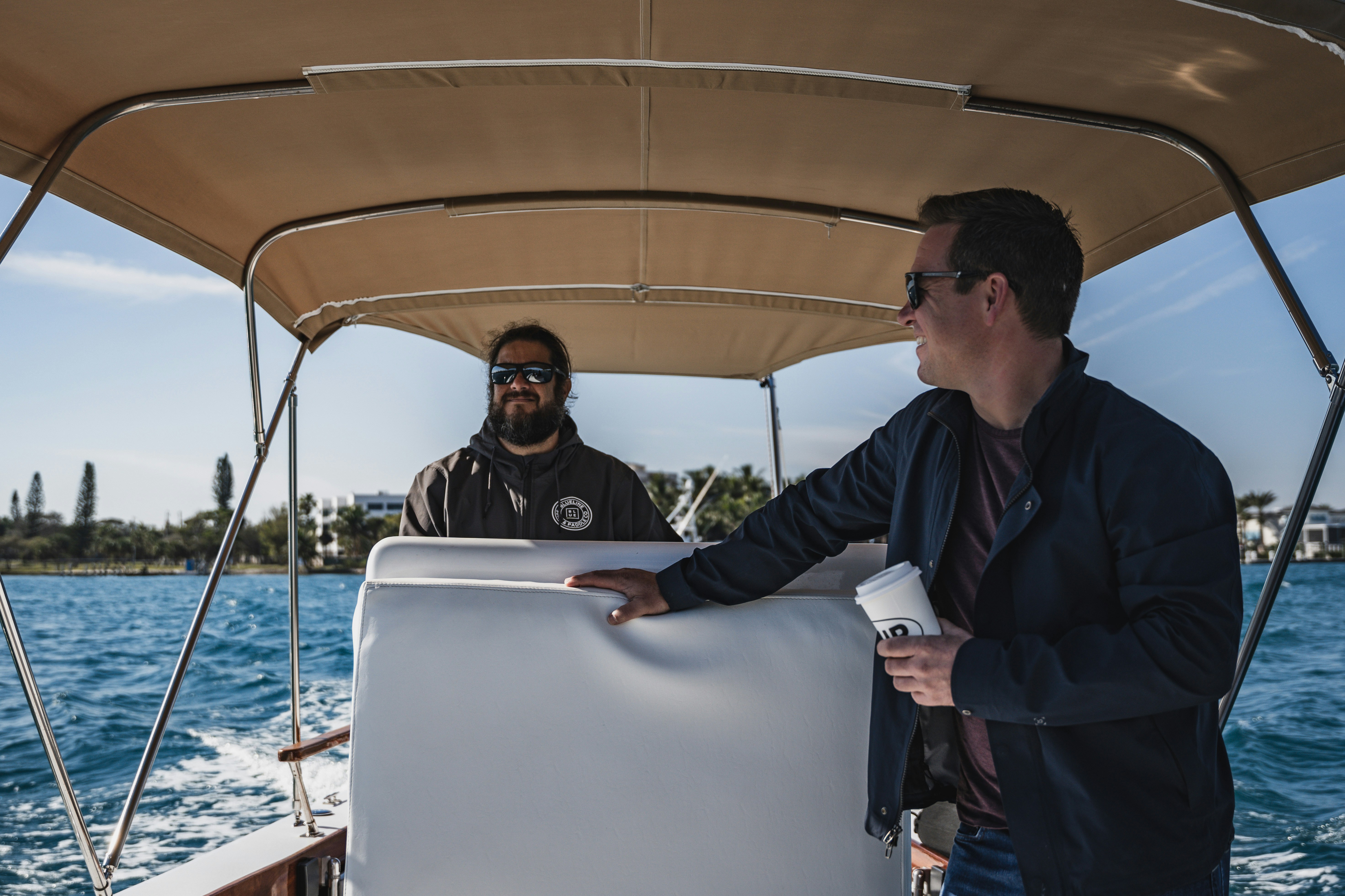 Two people enjoying a leisurely boat ride under a beige canopy with a clear blue sky and distant shoreline, one holding a coffee cup.