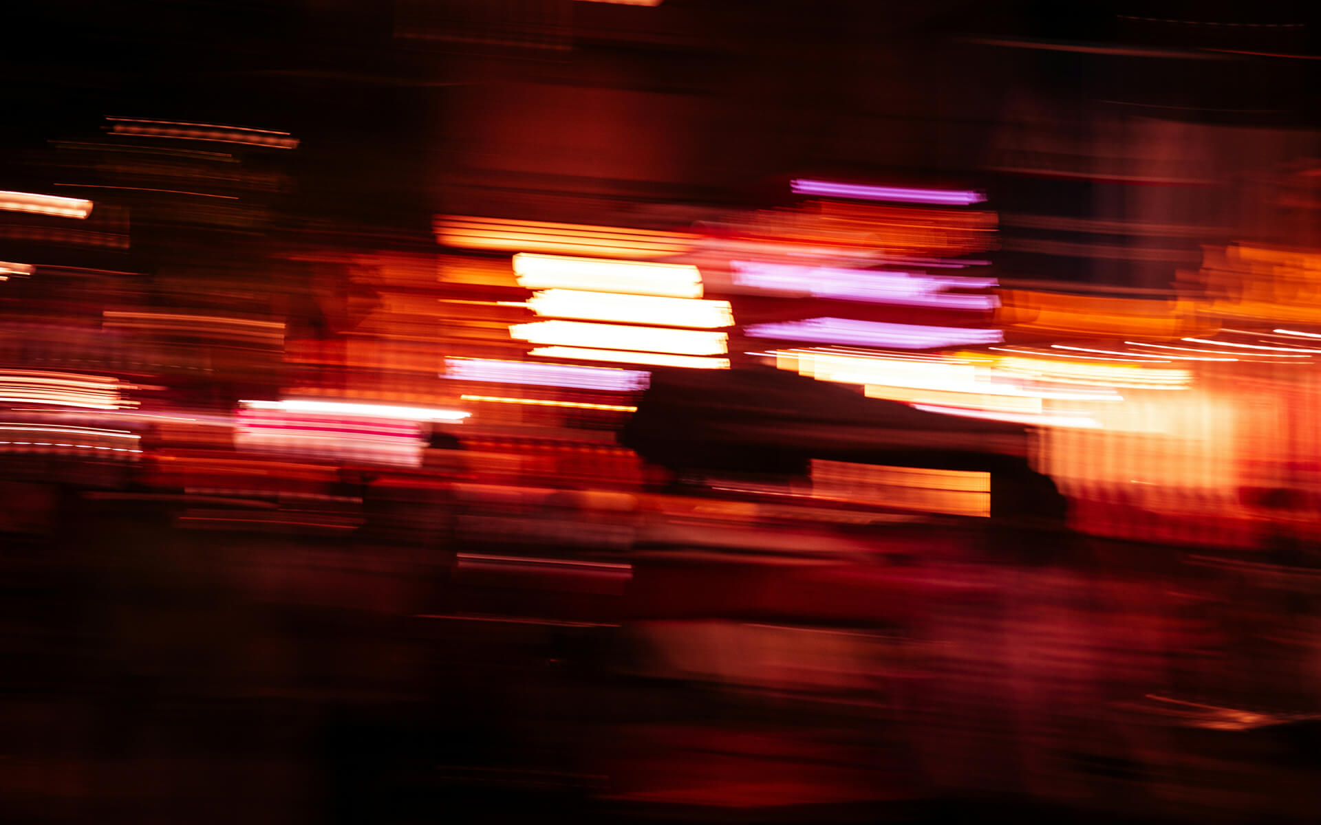 Abstract image of vibrant city lights in motion blur, with streaks of orange, red, pink, and purple cutting across the frame. The photo evokes a sense of speed and energy, capturing the dynamic atmosphere of a nighttime urban scene.