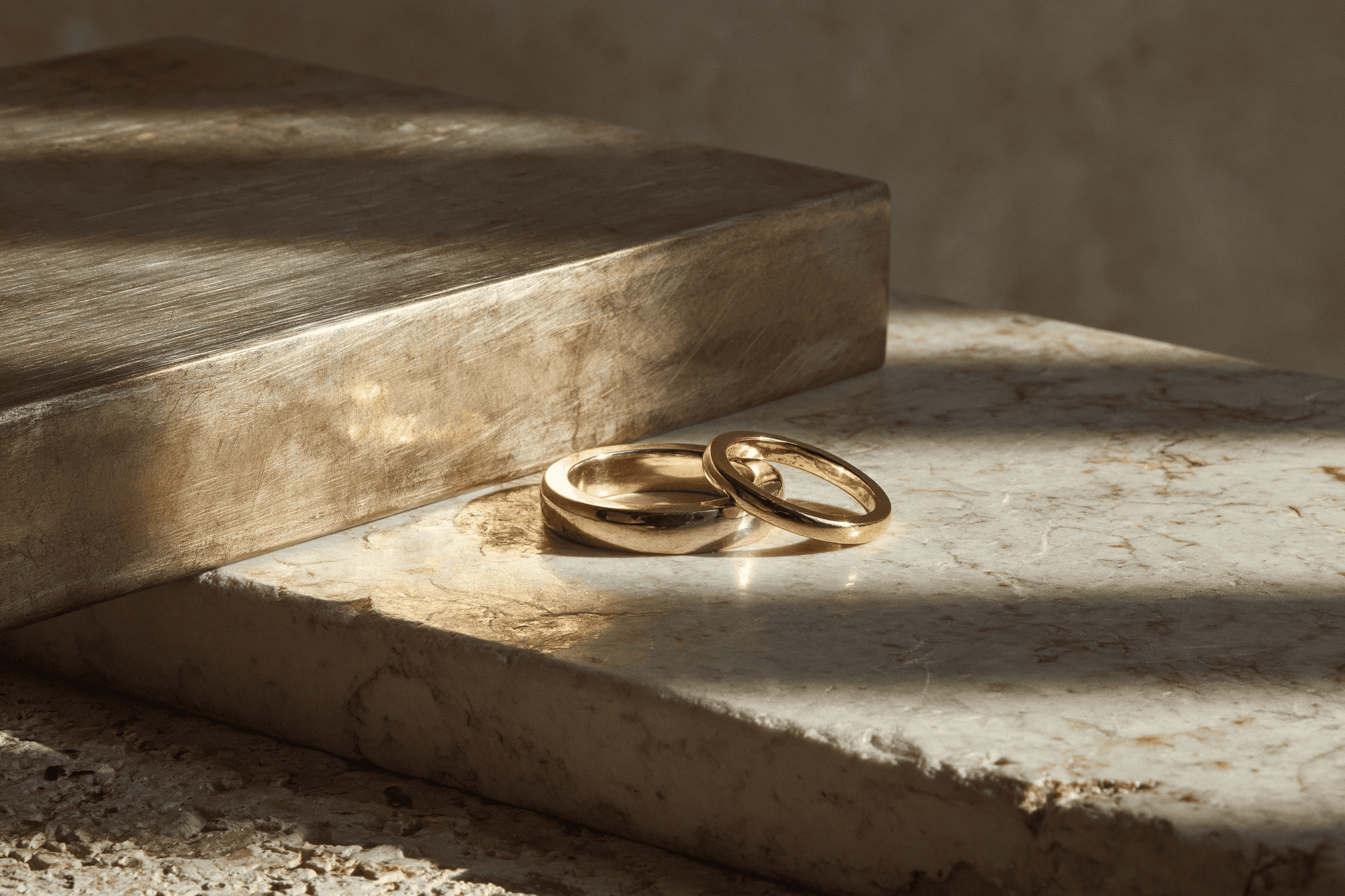 Two gold wedding rings rest on a sunlit marble step beside a wooden stair riser, with dramatic shadows cast across the textured surfaces.