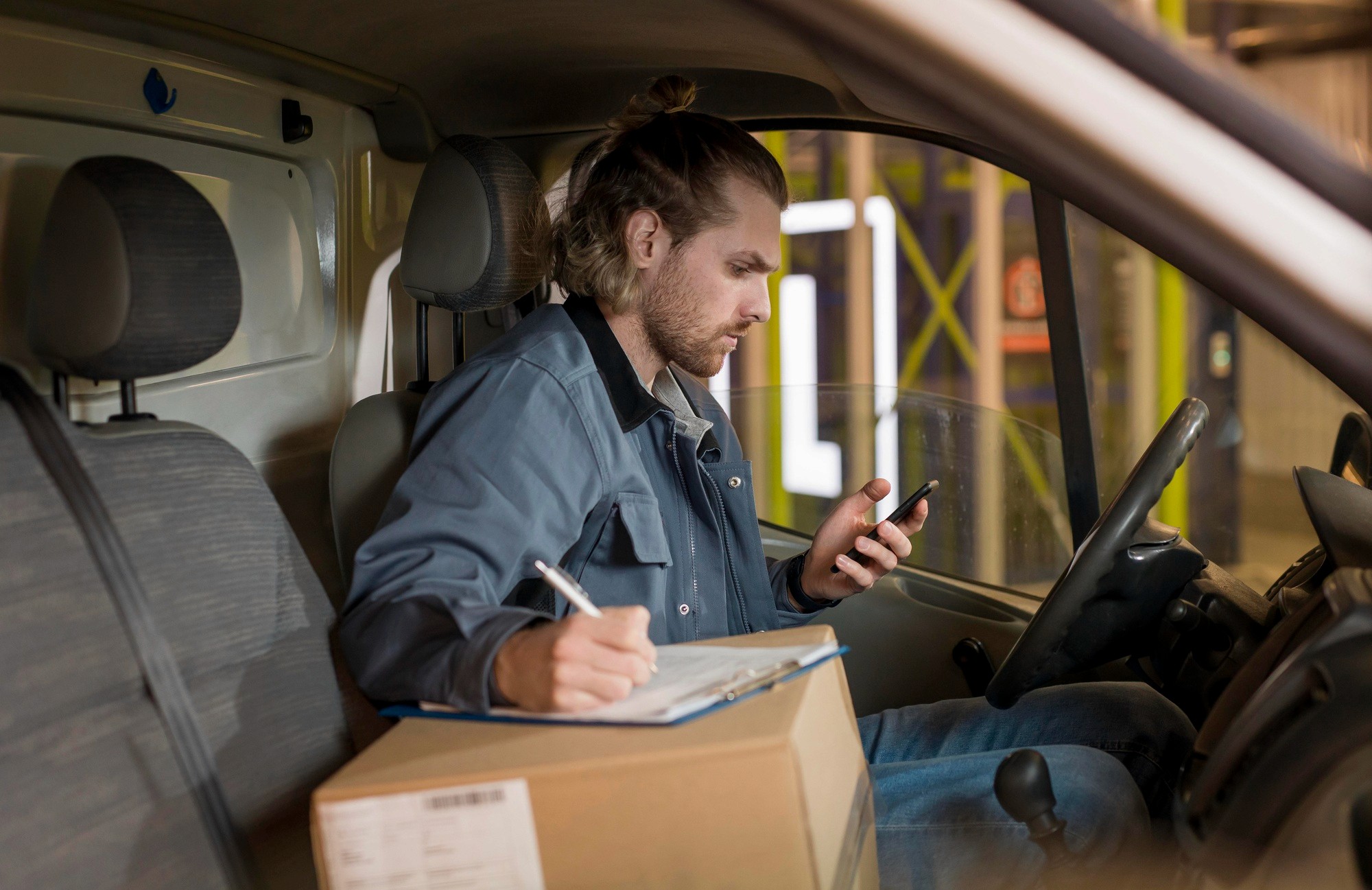A male delivery driver with a beard and a hair bun sits in the driver's seat of a delivery van or truck, wearing a blue-grey work jacket and jeans. He is looking down at his smartphone in his right hand while holding a pen in his left hand, writing on a clipboard that is resting on a brown cardboard box on his lap.