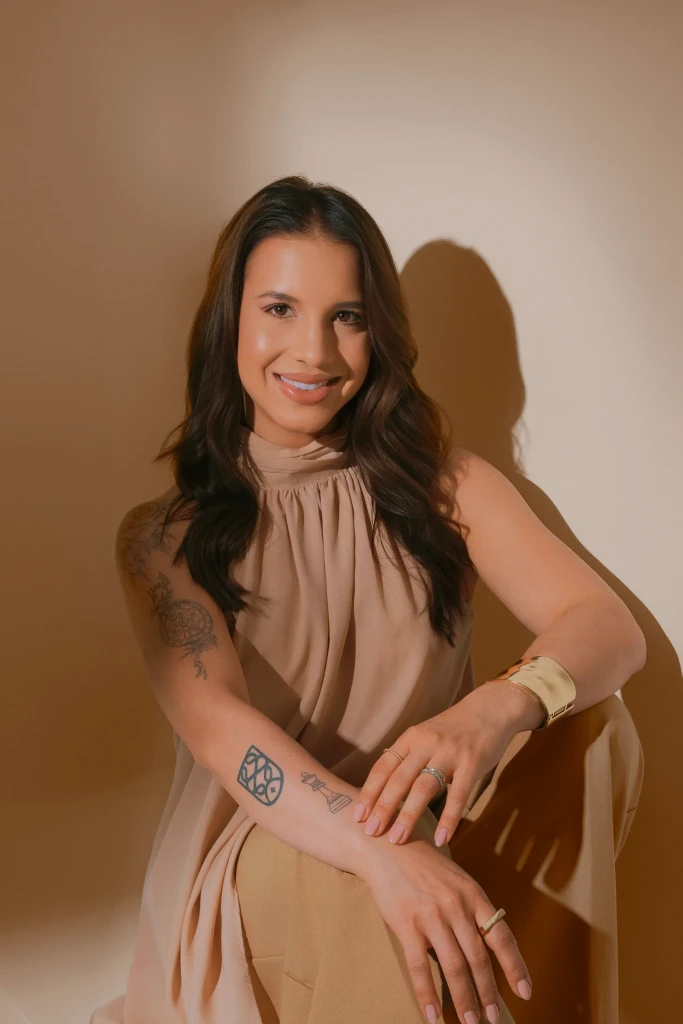 Isabela Ramos sentada sorrindo com tatuagens visíveis, pulseira dourada e look nude – foto profissional