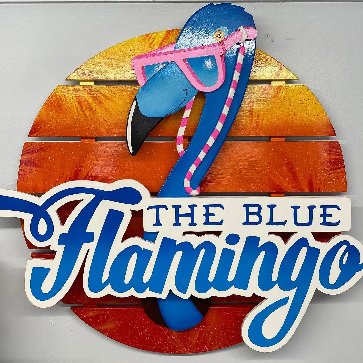 A vibrant sign featuring a stylized blue flamingo with pink sunglasses, set against a sunset background, prominently displays the text "The Blue Flamingo," capturing a fun and colorful theme in Bradenton, FL.