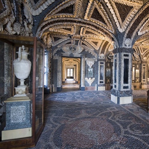 Ornate room with intricate shell and stone decorations on walls and ceiling; a white urn is displayed in a glass case.