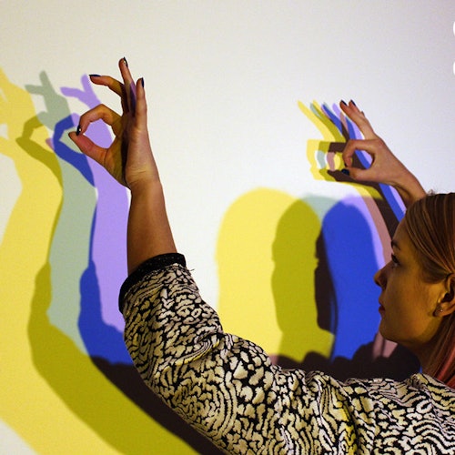 一个人在白墙上用彩色的手影表演,重叠的灯光投影呈现出黄色、蓝色和紫色的视觉效果。