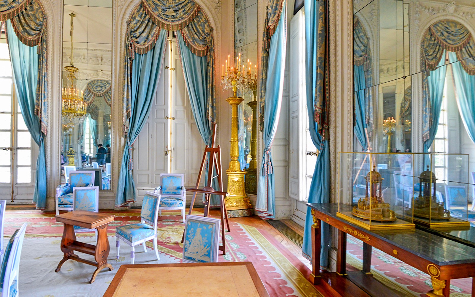 Ruang Grand Trianon dengan tirai biru yang berhias indah, tempat lilin berlapis emas, dan furnitur yang elegan.