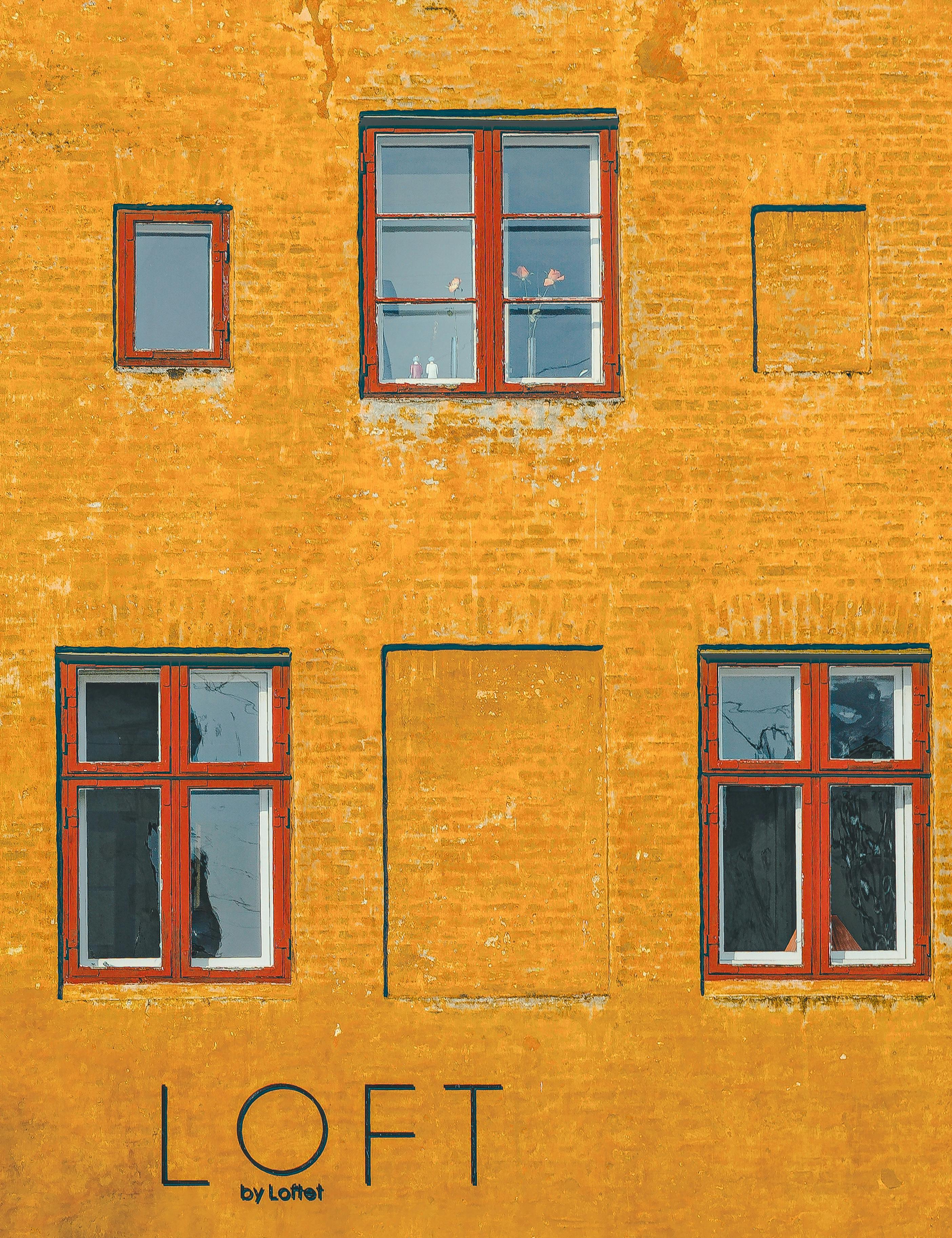 A textured yellow brick wall with three red-framed windows. The text "LOFT by Loftet" is written at the bottom.