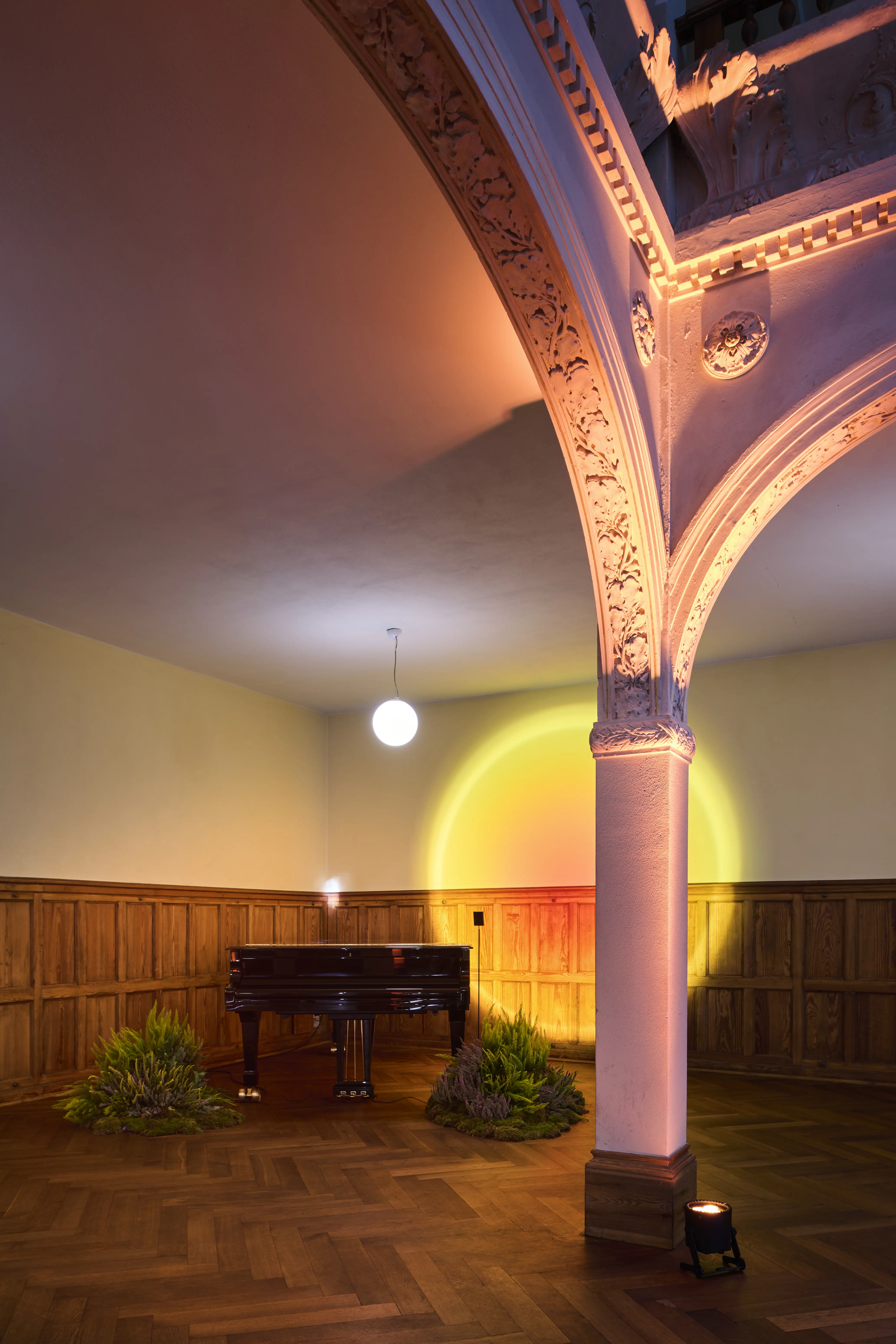 Historic hall corner with grand piano, ornate arch, plants and a sunset light circle on the wall.
