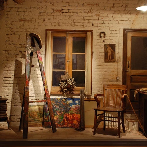 一个艺术家的工作室，里面有一张沾满油漆的画架，装裱好的画作，一把木椅和一堵砖墙上的窗户。