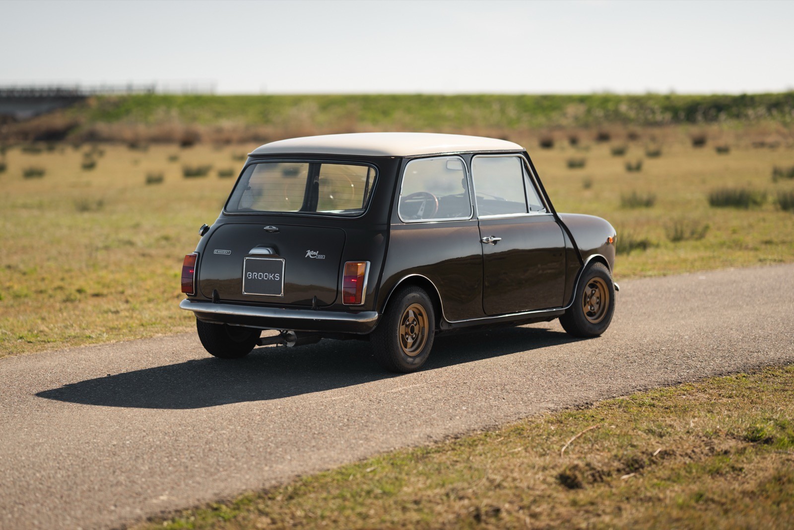 A small vintage car parked on a rural road, with open landscapes in the background under clear blue skies.