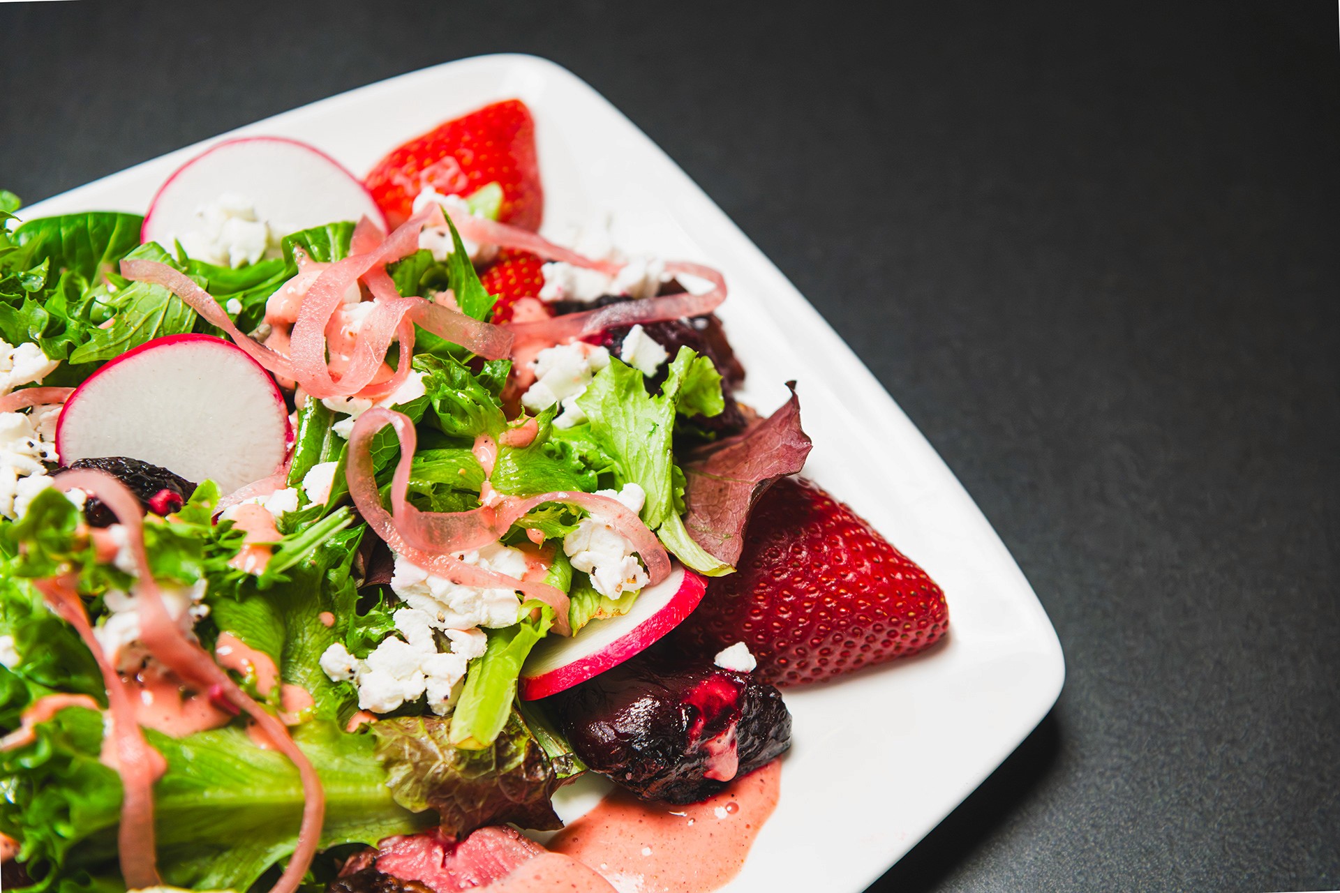 A crisp summer salad on a square white plate, featuring mixed baby greens, sliced radishes, strawberries, and crumbled feta cheese with a light dressing.