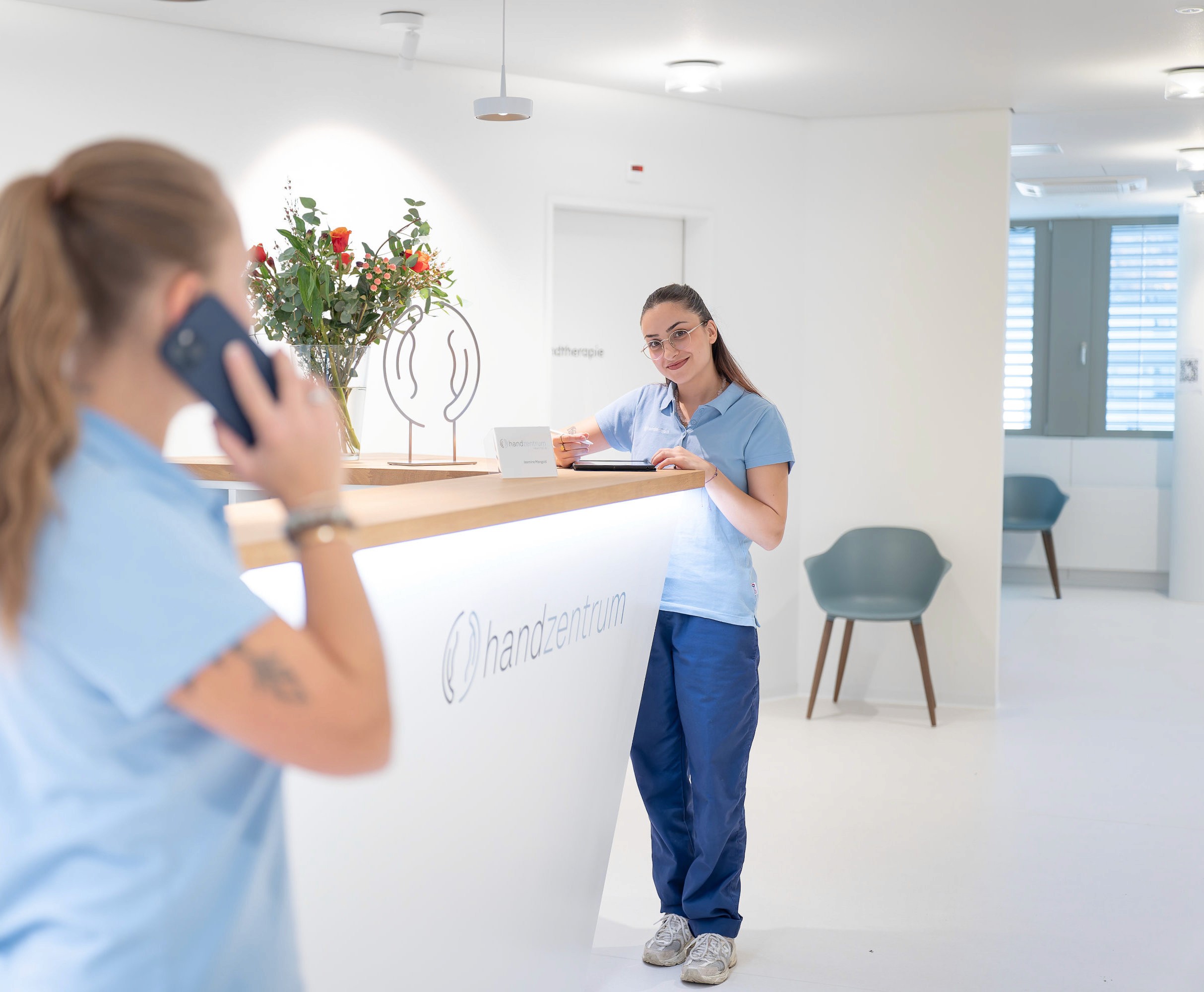 Zwei Frauen in hellblauen Uniformen stehen am hellen Empfang des Handzentrums Pratteln. Die eine nimmt Terminvereinbarungen am Telefon entgegen, während die andere auf einem Klemmbrett schreibt. Eine Vase mit Blumen schmückt den Schreibtisch, im Hintergrund stehen Stühle.