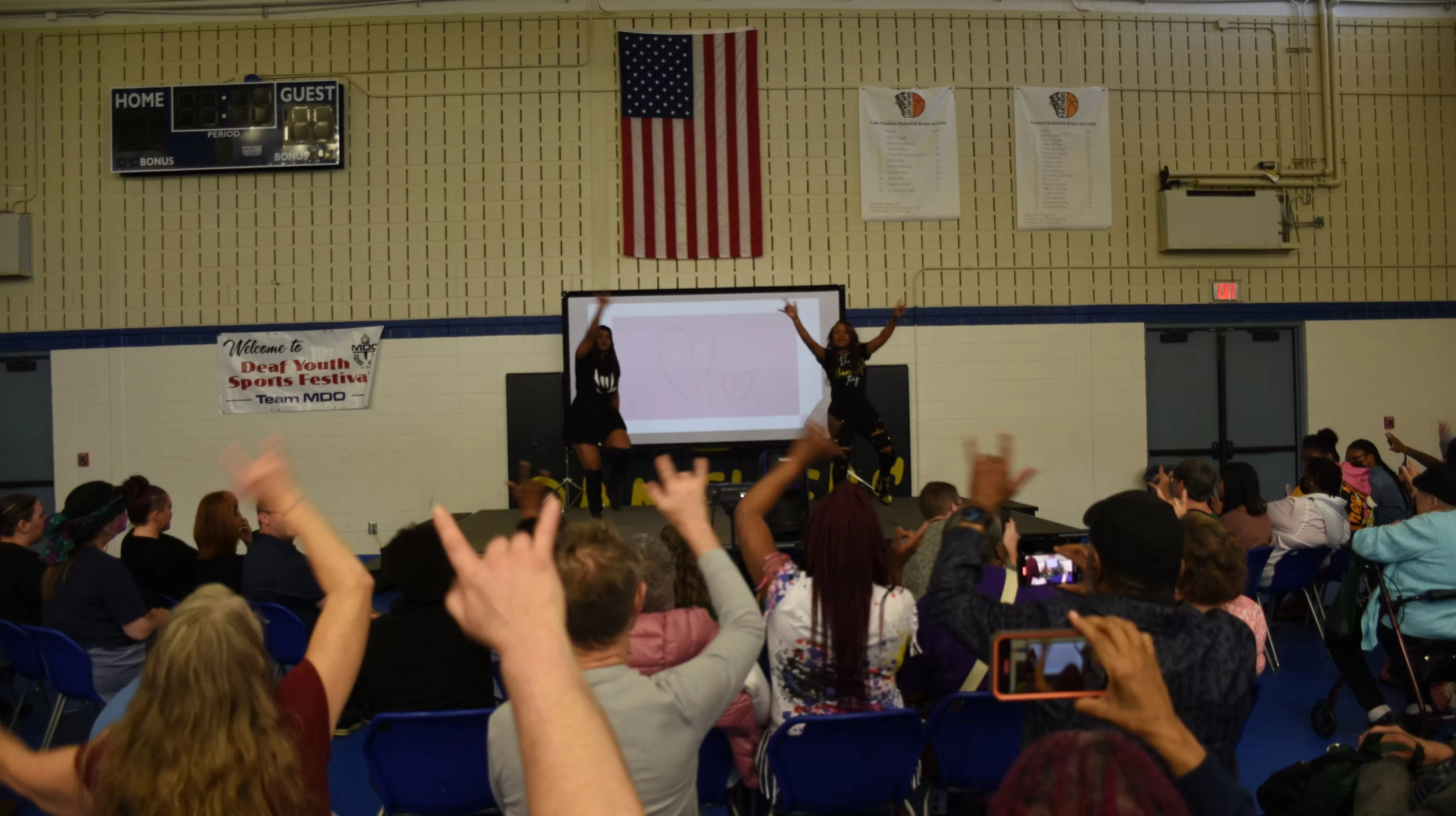 Two performers on stage in front of an audience. The audience is cheering and raising their hands. A large screen is behind the performers.