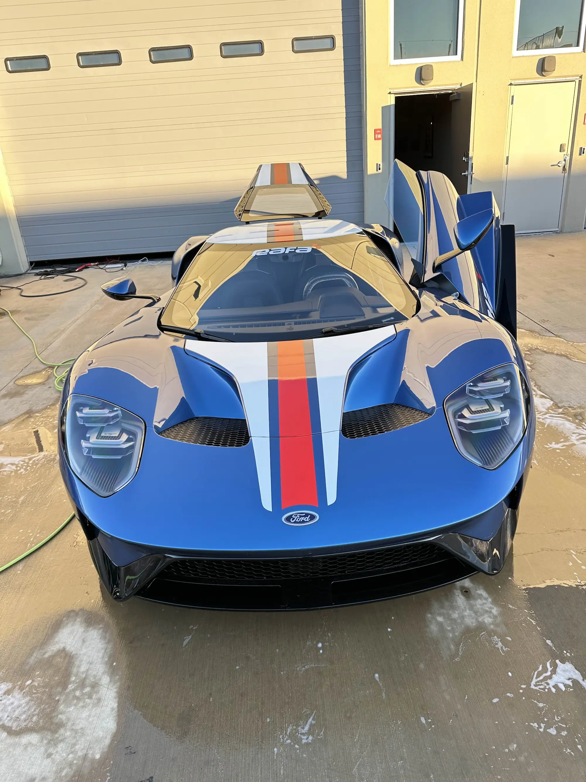 Front view of a blue Ford GT supercar with white and orange racing stripes and butterfly doors open at a professional Houston car detailing shop.