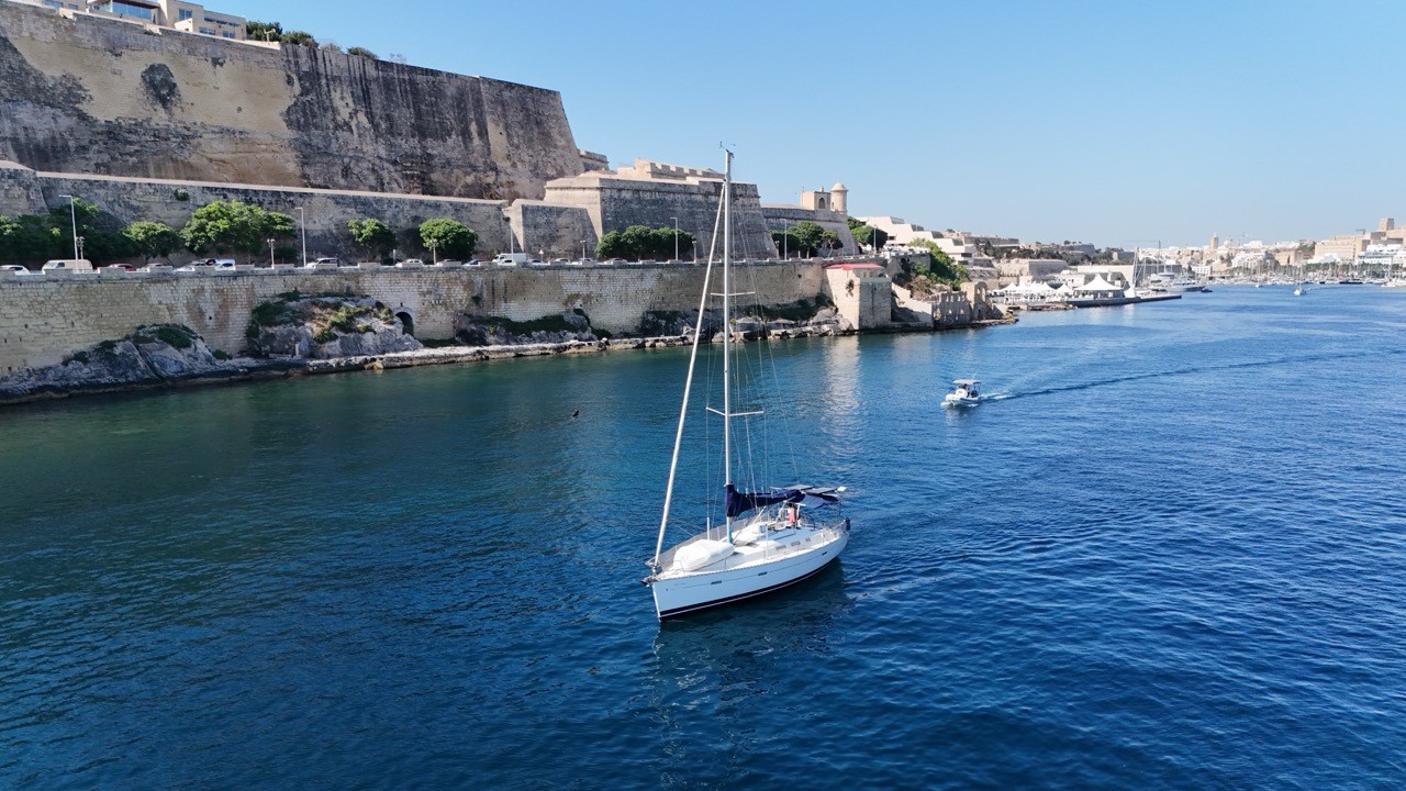 Aerial drone view of a white Beneteau Oceanis Clipper 373 sailboat motoring through the Grand Harbour, Malta, with the imposing limestone bastions of Valletta's fortified walls rising steeply to the left. A small motorboat follows in the distance, with a marina visible further along the harbour.