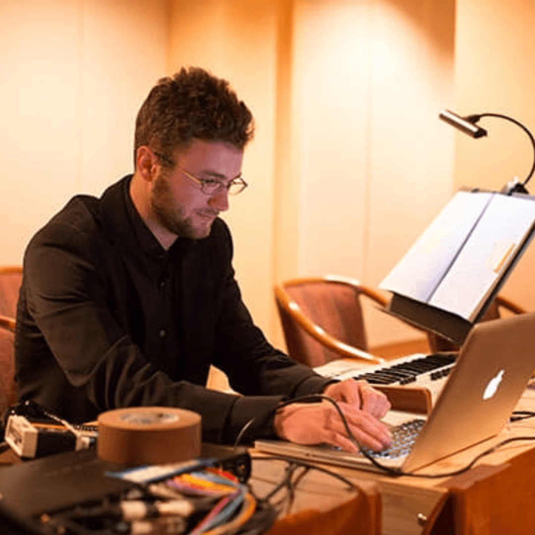 a man sits at a computer surrounded by tape, wires, sheet music, and soft lights