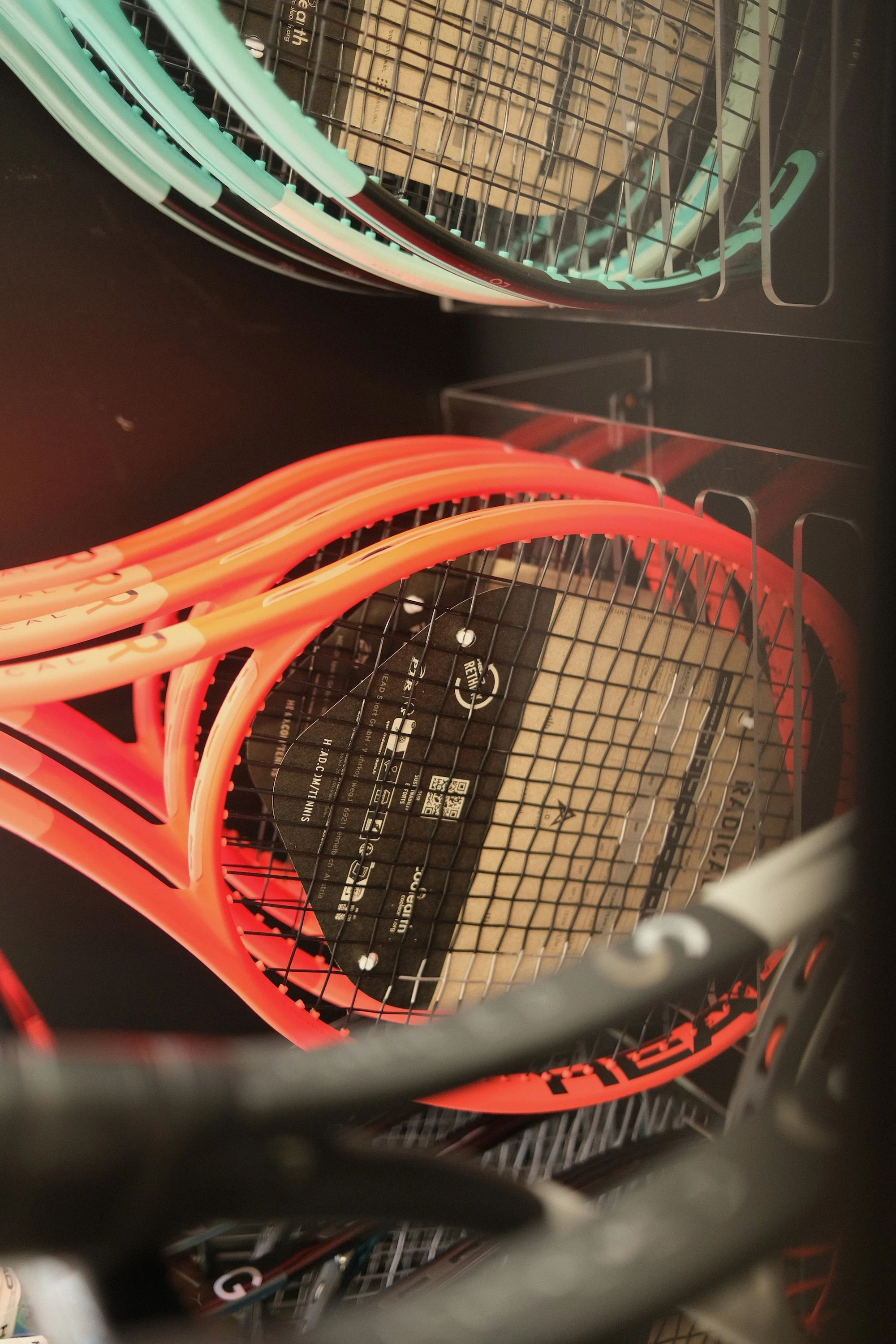 Eine Nahaufnahme eines Schlägerregals mit mehreren HEAD Tennisschlägern, darunter Modelle der Radical- und Gravity-Serie. Die Schläger haben leuchtend orangefarbene und türkisfarbene Rahmen und sind unbespannt. Die Rahmen sind ordentlich in einem Metallhalter platziert, während im Hintergrund weitere Schläger sichtbar sind.