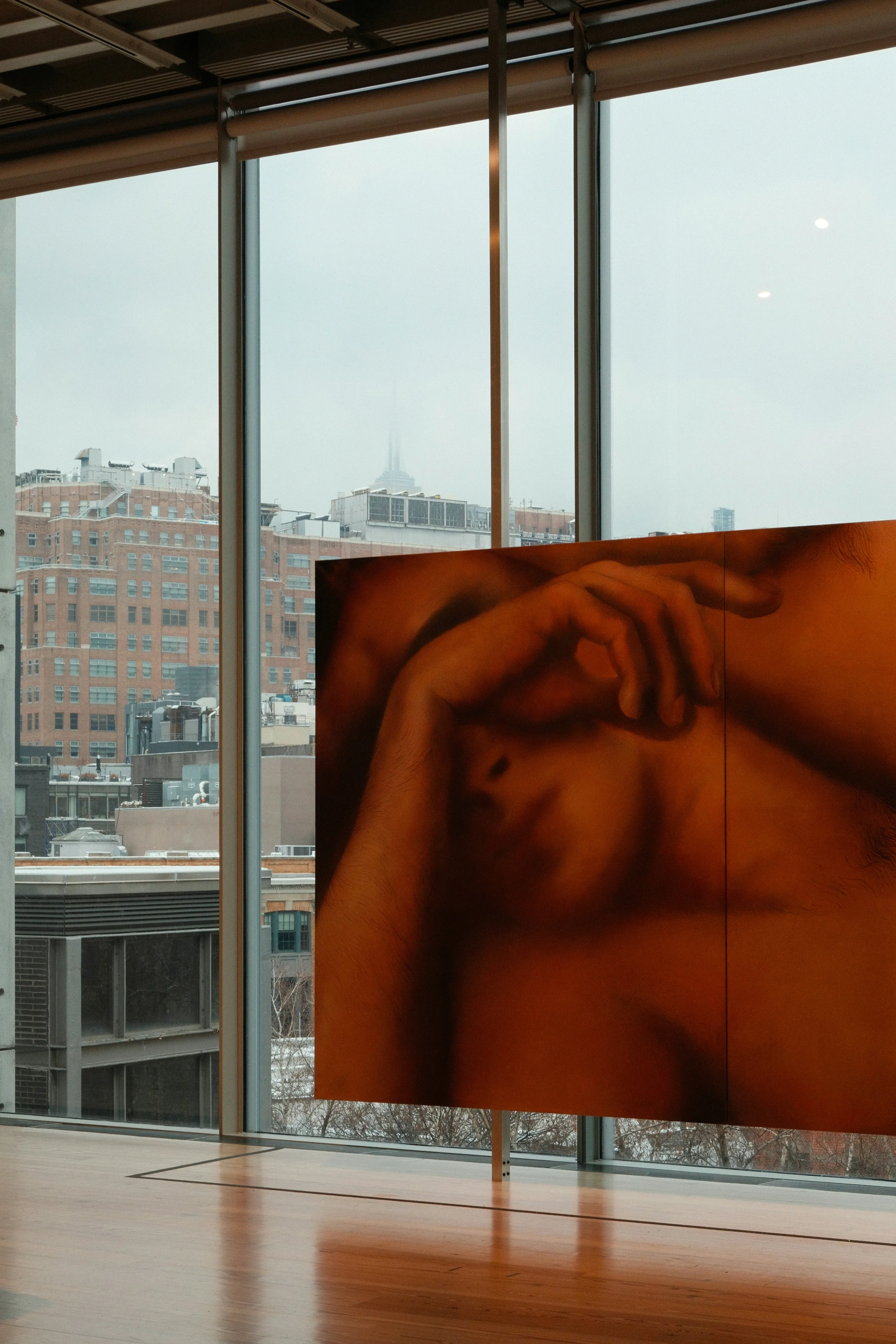 Artwork of a woman shielding her eyes with her arm sits in front of a floor to ceiling glass window at the Whitney Museum of American Art in New York City.