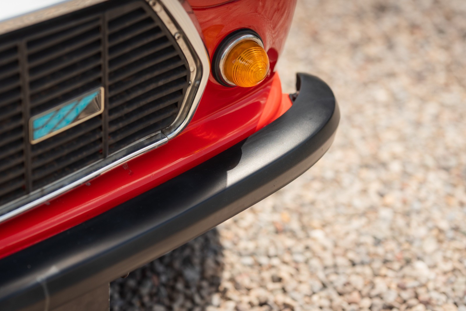 Close-up of a car's front bumper, showcasing a shiny grille and orange turn signal on a gravel surface.