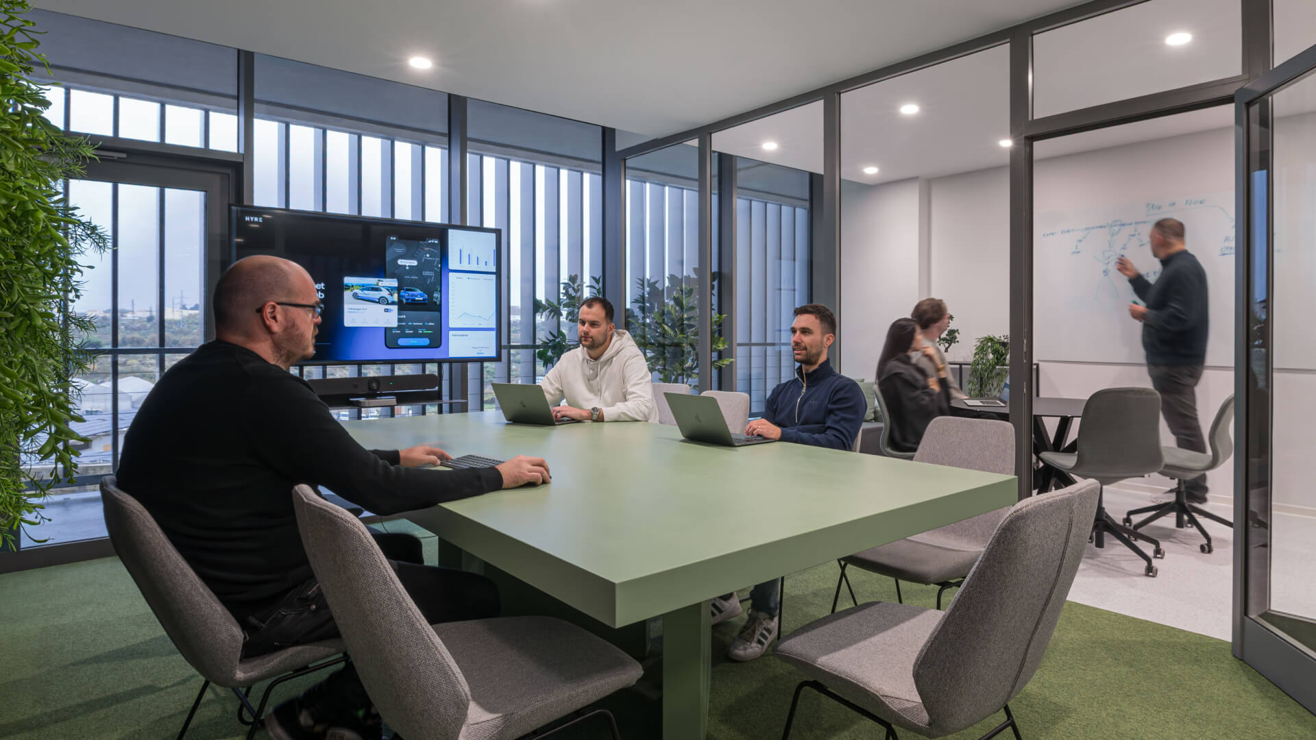 A modern office meeting space where a product development team collaborates around a large green conference table with laptops. On the left wall, a large digital screen displays a mobile application interface with data charts. In the background, another team member writes a diagram on a whiteboard inside an adjacent glass-walled meeting room, showcasing a dynamic, multi-team workshop environment.