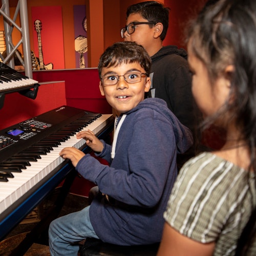 A young boy plays a keyboard with two other children nearby, all smiling, in a room with colorful walls and musical artwork.