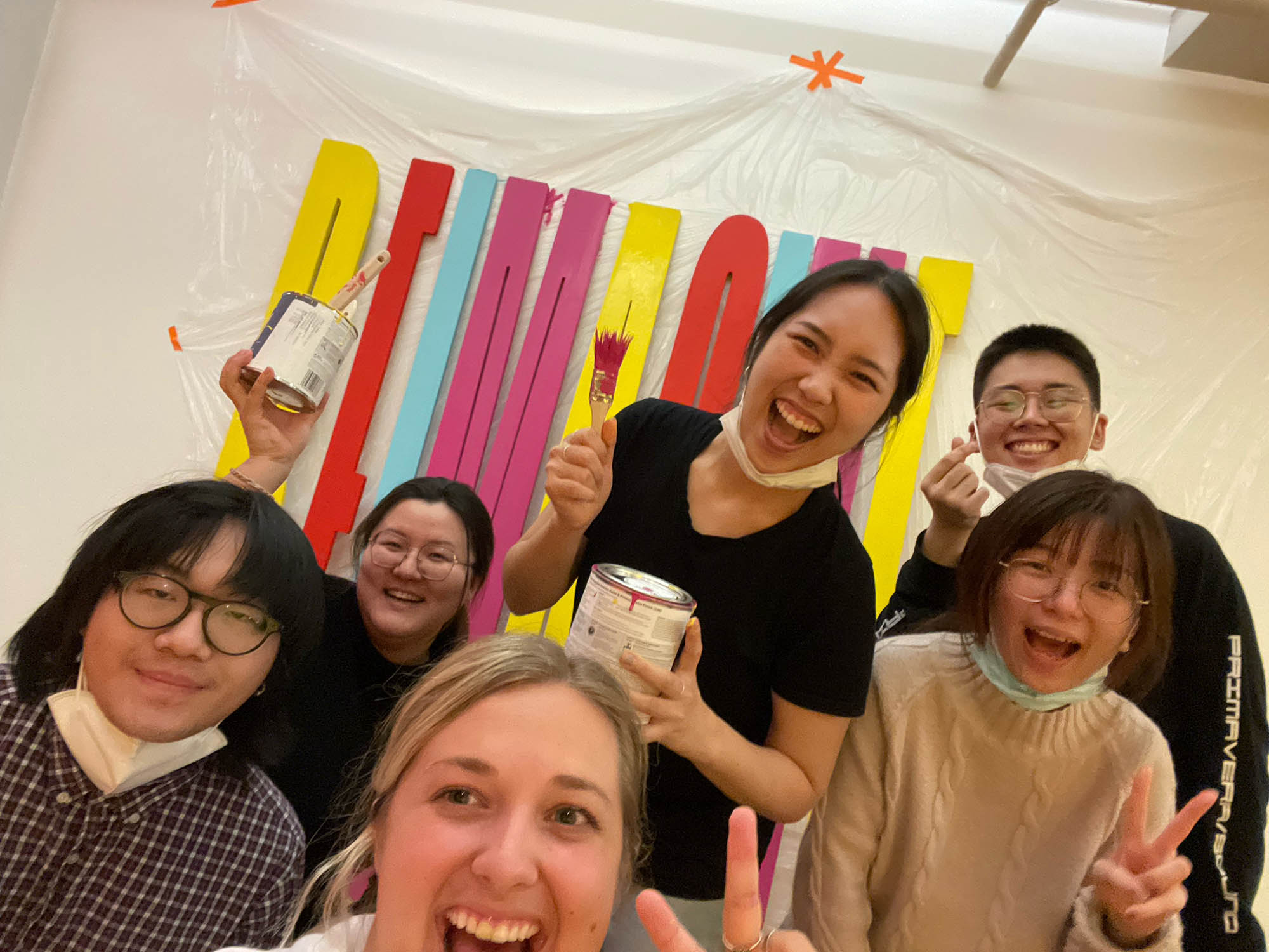 A group of smiling students with paint buckets and brushes in their hands. Also behind them there is a colorful 3d text on a wall that says: REIMAGINE.