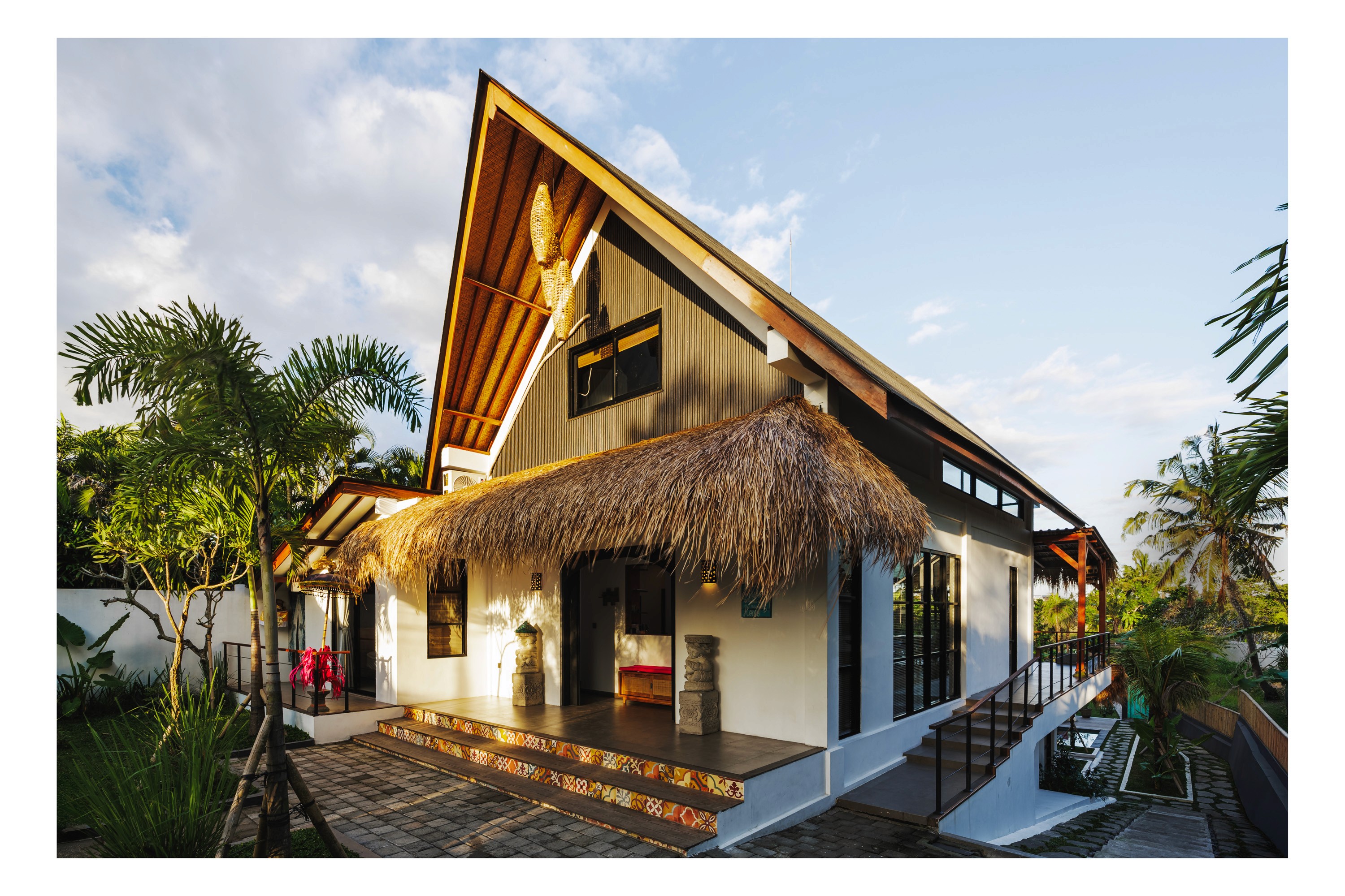 Modern tropical villa with thatched awning, pitched roof, and palm-lined entrance at golden hour.