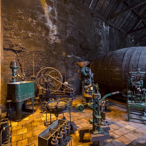 An old workshop with vintage machinery, large wooden barrel, stone walls, and a brick floor.