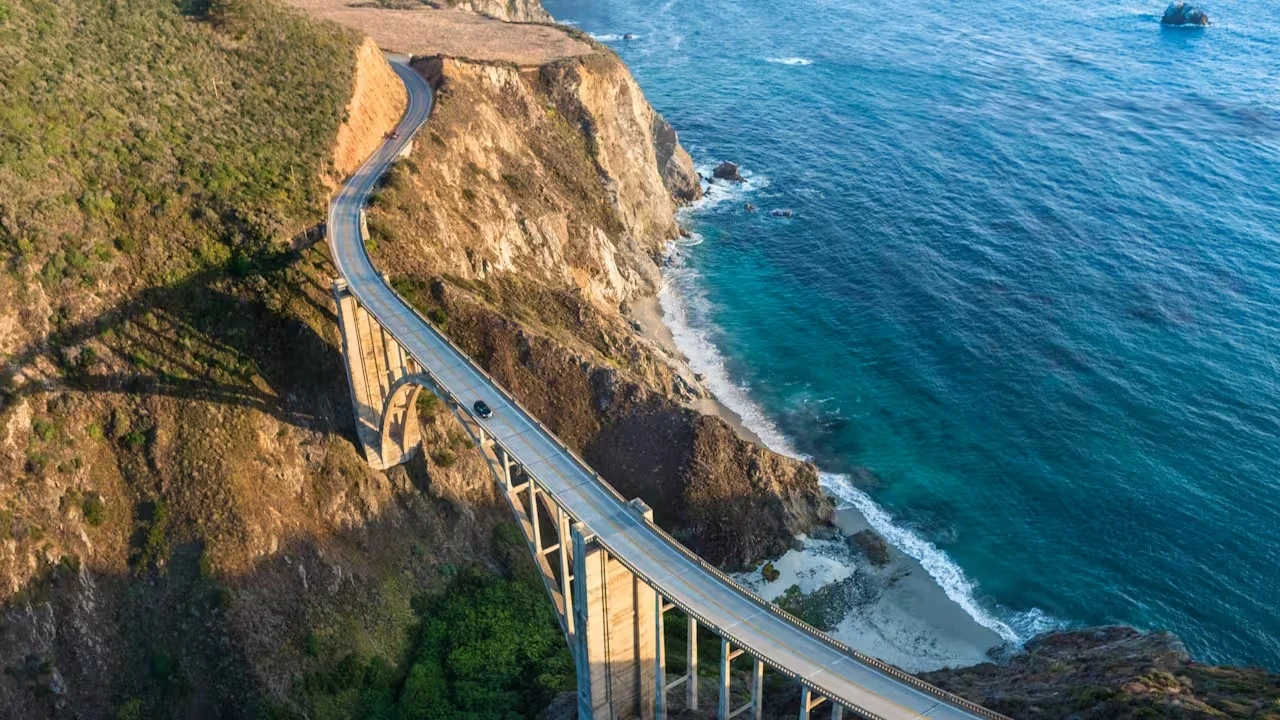 Aerial view of a curved concrete bridge hugging a rocky coastal cliff above bright turquoise water.