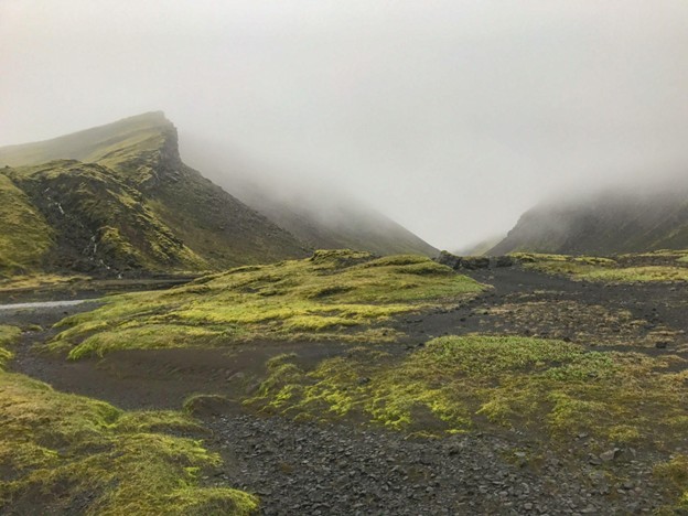 The picture shows the Eldgjá canyon in Katla Geopark, in the Highlands of south Iceland from the air. The Eldgjá volcanic fissure eruption that formed this canyon was the largest on earth in the past 1000 years, and the canyon itself stretches 40 kilometers in length. There is a long, lush green canyon, and black and red lava beds and sands. A waterfall pours down the side of the mossy canyon.