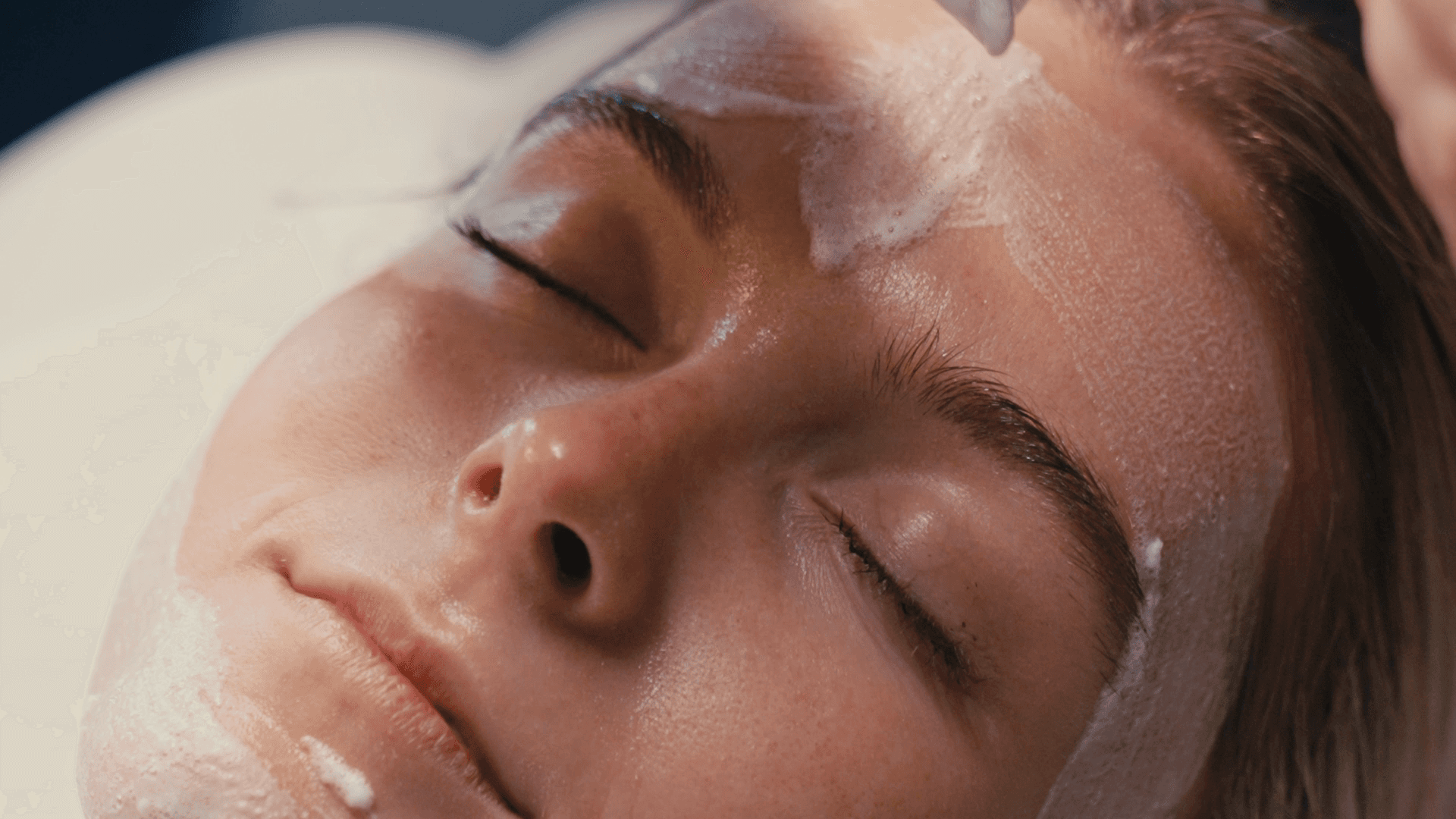 A woman lying down with a towel wrapped around her head, her face glistening with facial oil. Her eyes are closed, and she appears to be enjoying a spa treatment in a peaceful setting.