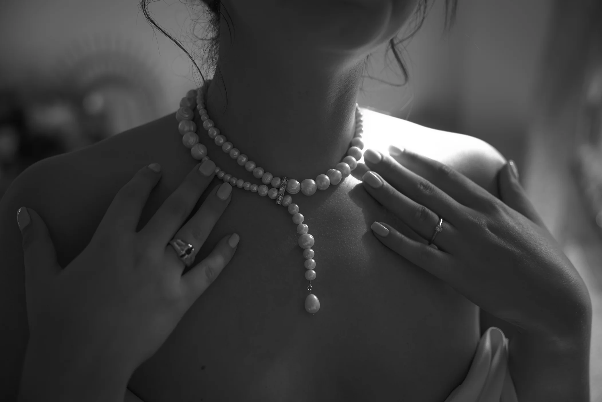 Black and white close-up of a bride's neck and shoulders adorned with a long pearl necklace, her hand with an engagement ring resting on her shoulder