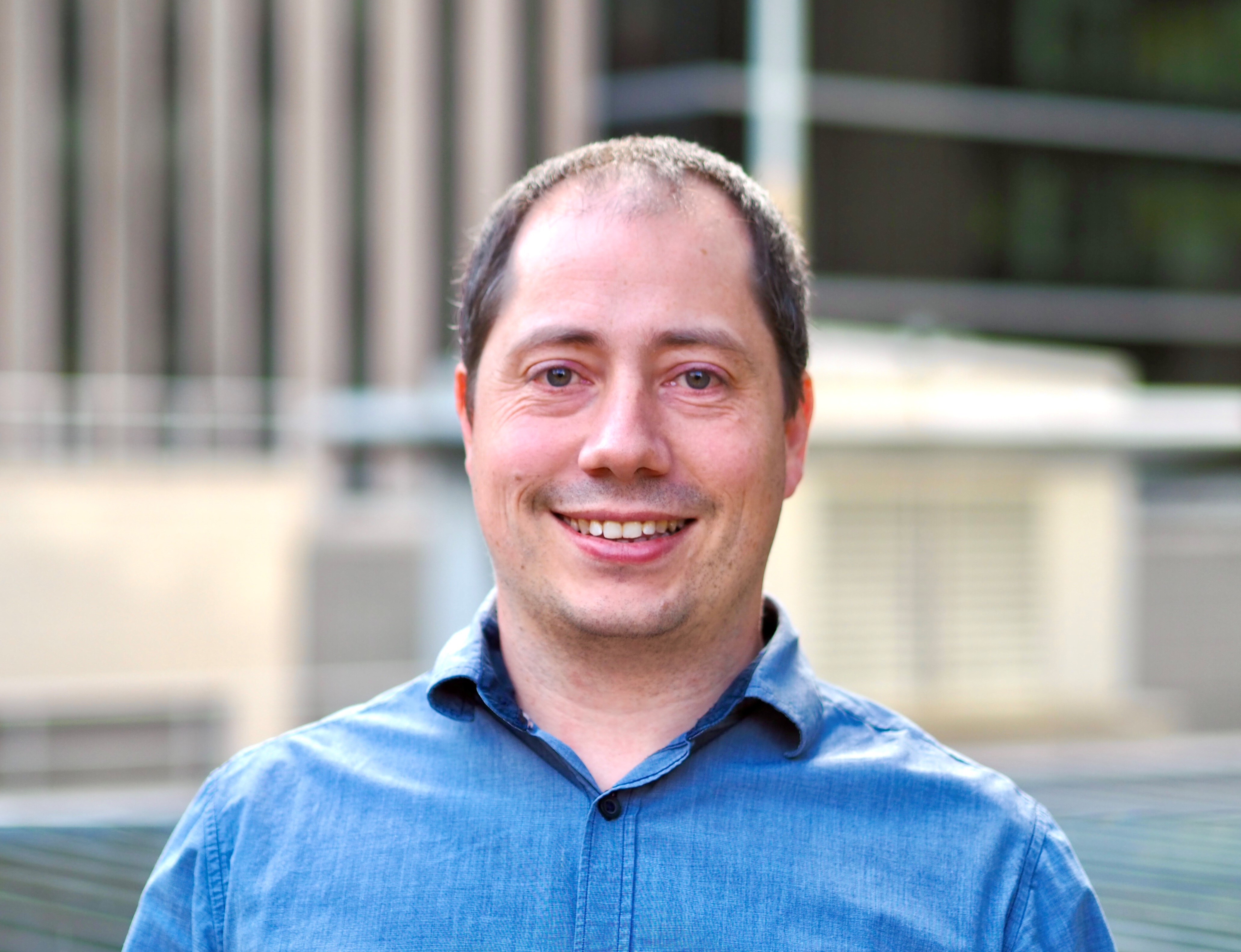 A woman with long, dark hair and a slight smile poses in front of a bright yellow background. She is wearing a black top and a necklace.