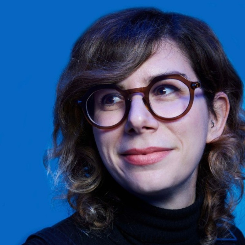 A confident person with short, styled hair and a leather jacket poses against a dark backdrop.