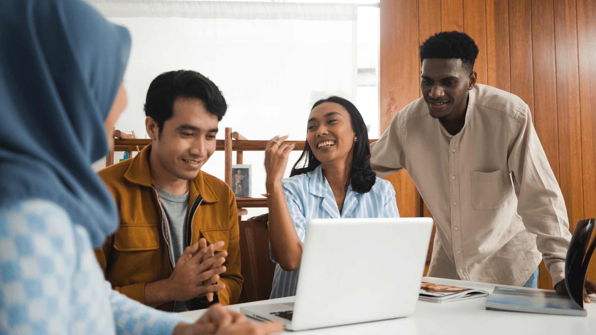 Three people, two men and one woman, sitting together and smiling while working on a laptop. They appear to be engaged in a collaborative work or discussion.
