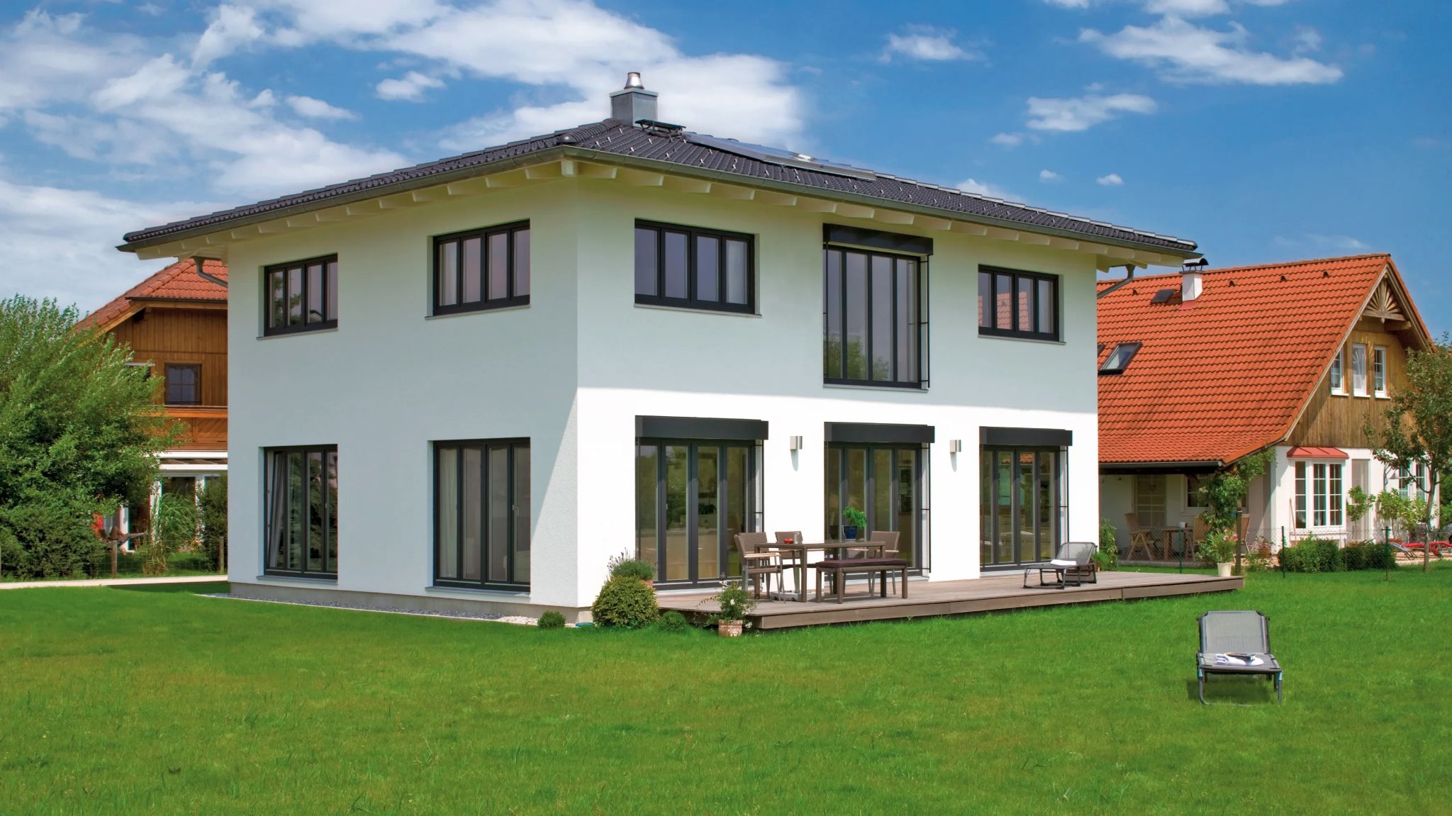 Modernes weißes Haus mit großen Fenstern, Solaranlage auf dem Dach und Terrasse mit Gartenmöbeln, daneben ein Haus mit rotem Ziegeldach.