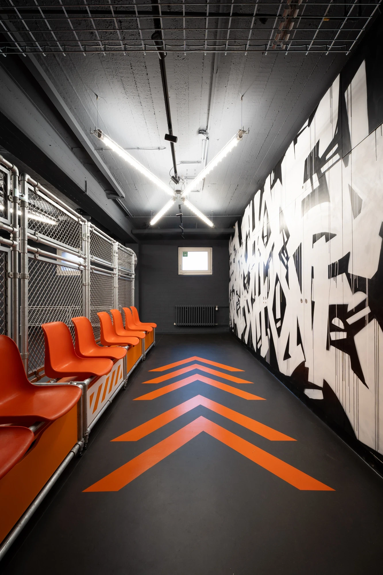 Industrial-style sports interior with orange plastic seating mounted on a chain-link cage, monochrome graffiti mural and orange chevron pattern painted on the floor.