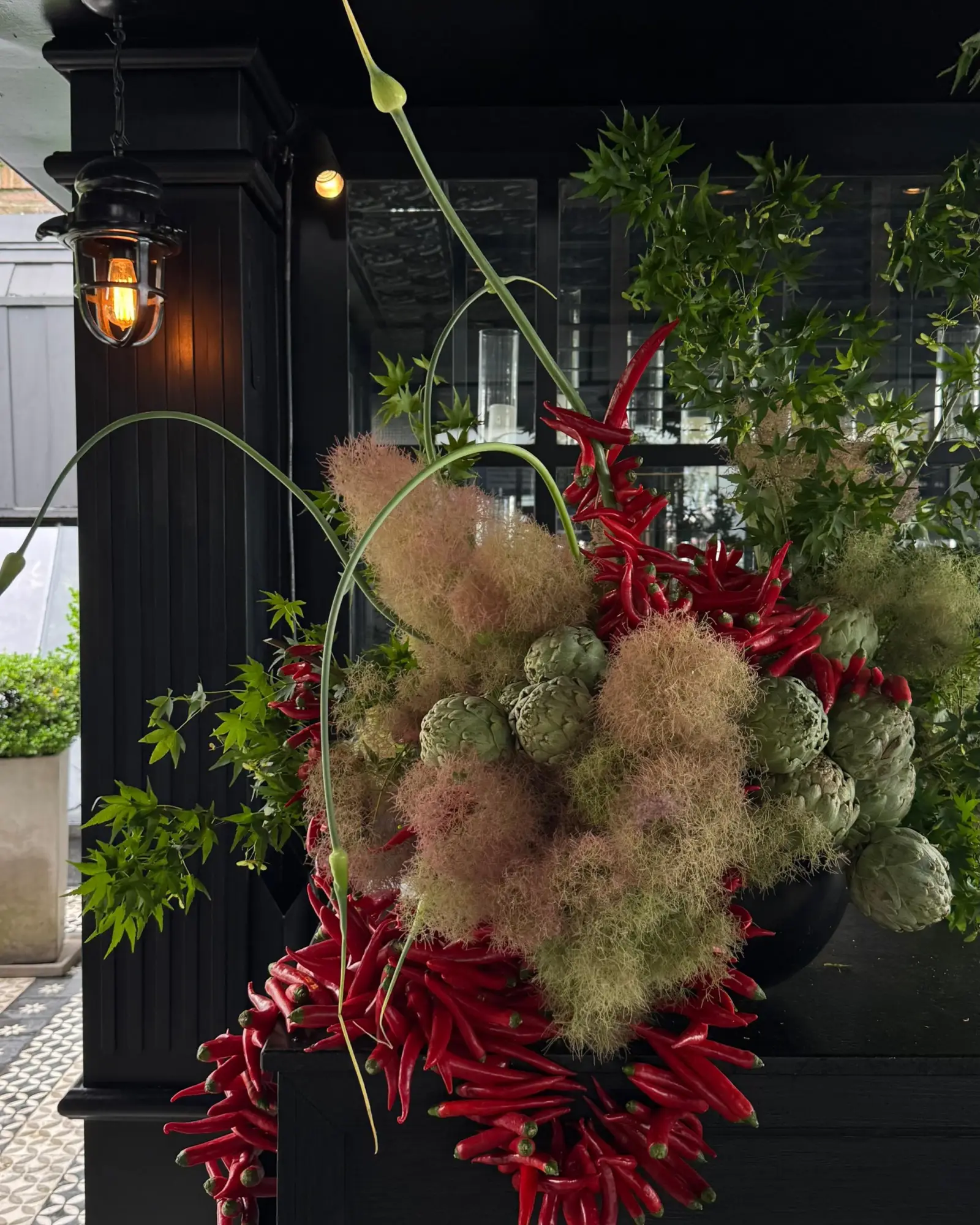 Composición floral y culinaria con alcauciles frescos, smoke bush rosado y cascada de ajíes rojos picantes en jarrón negro.