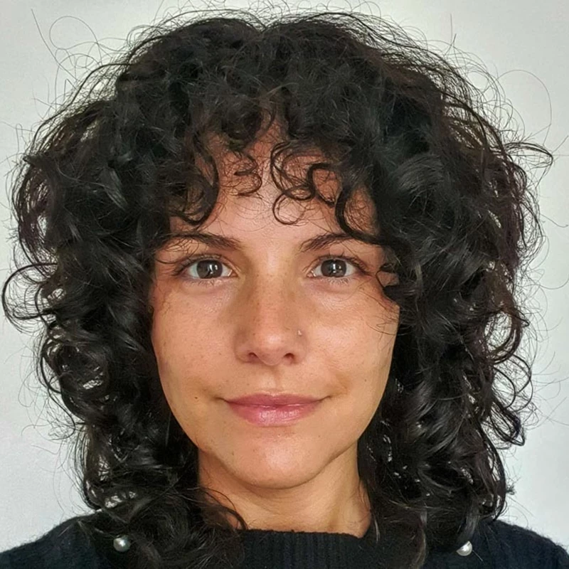 Person with short, voluminous curly dark hair wearing a black top with small decorative details, standing against a plain light background.