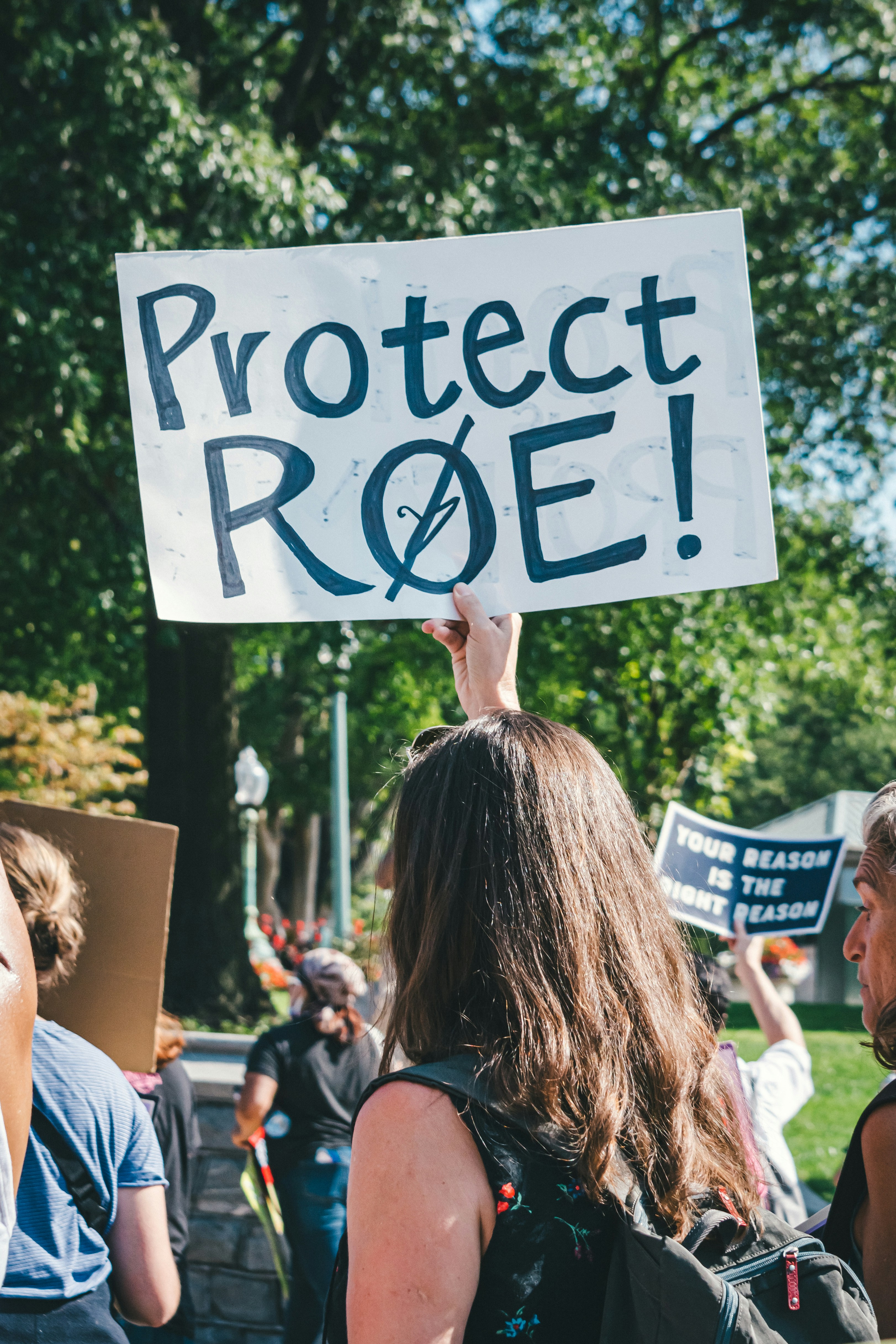 A protestor holding up a sign with one hand that reads, "Protect Roe" and has a slash through the "o" with the slash also going through a coat hanger