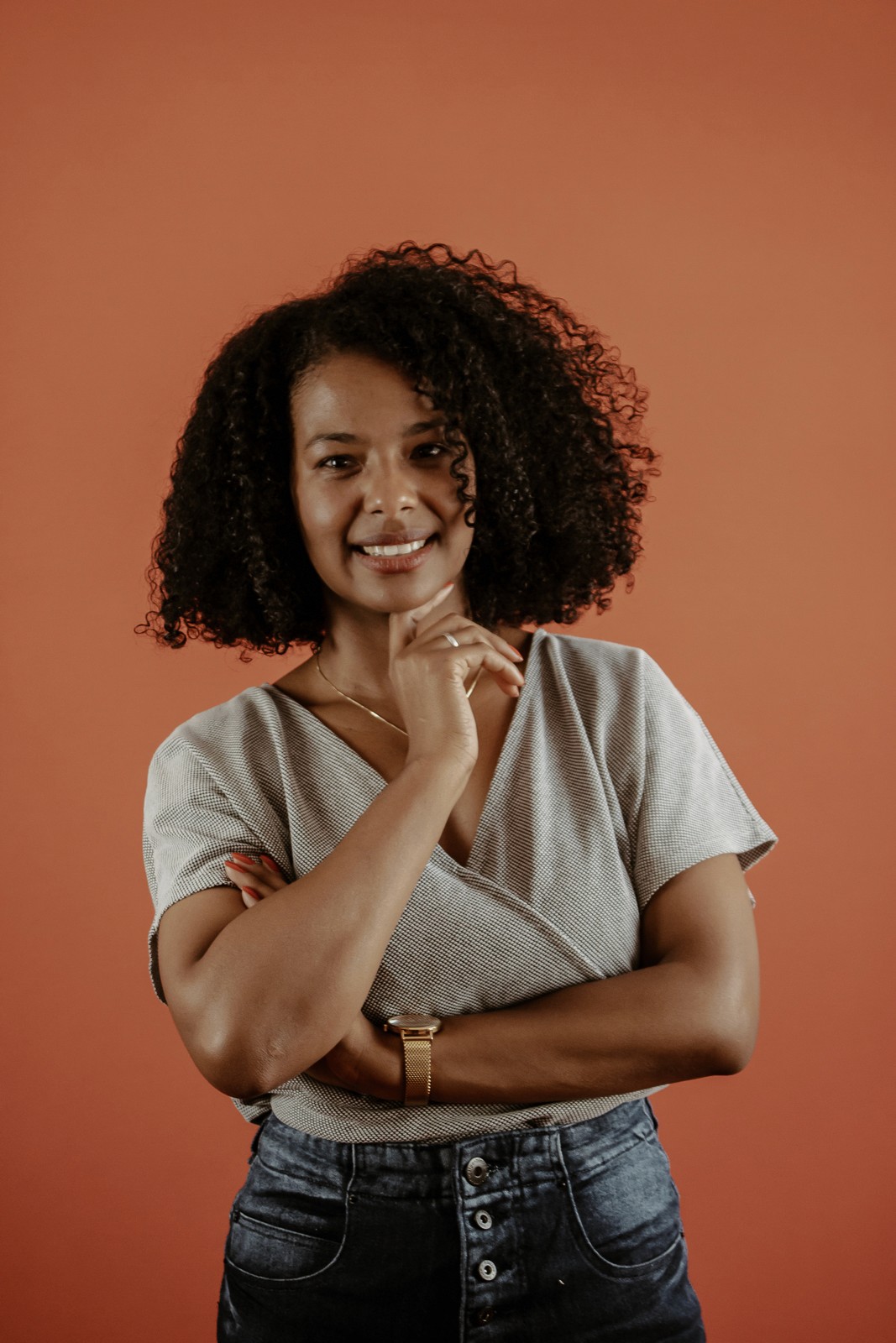 Mulher negra de cabelos médios acima o ombro, sorrindo para a camera com a mão no queixo, camisa bege e fundo laranja