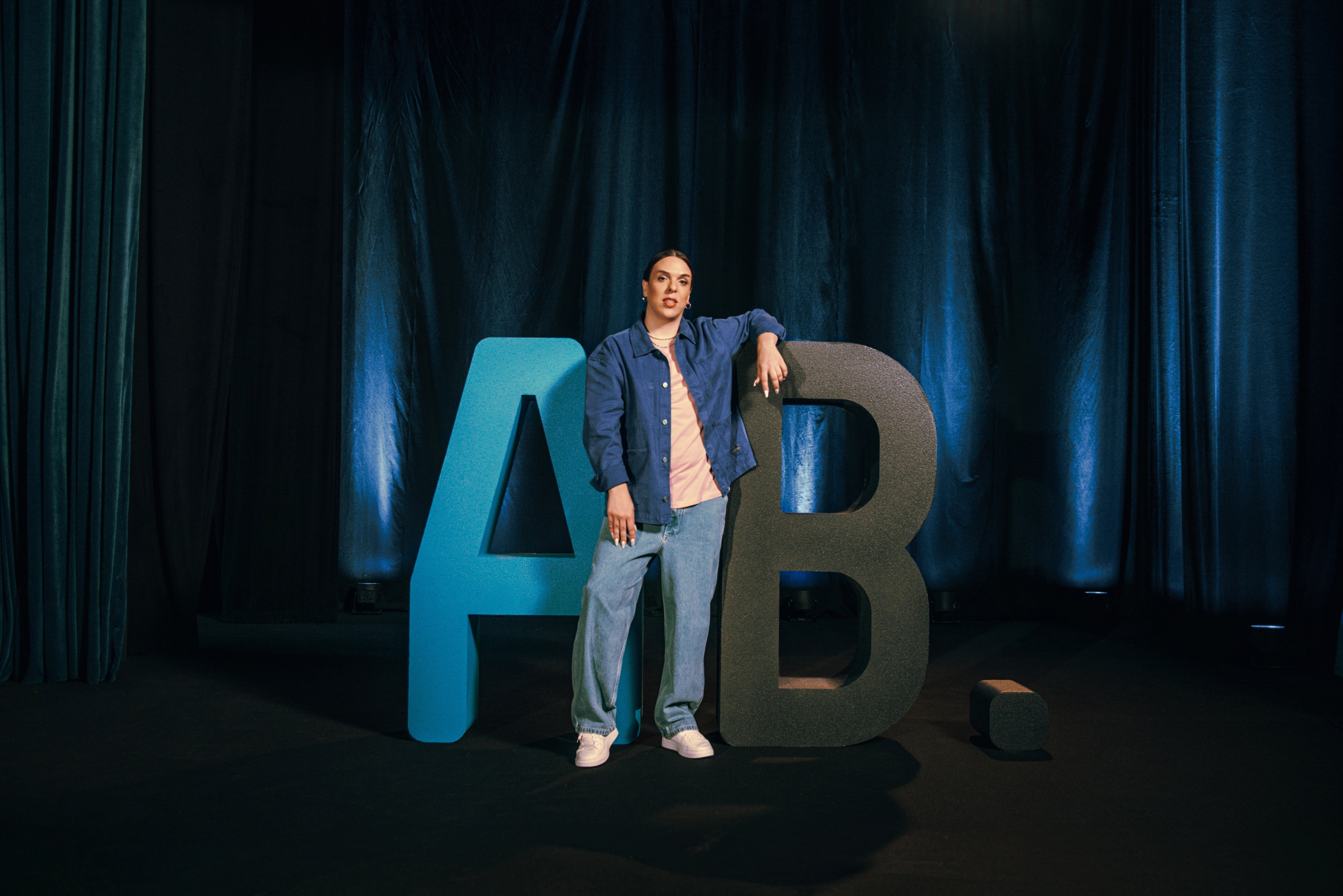 Kiko is Hot in a denim jacket leaning on the giant AB letters on a dark stage